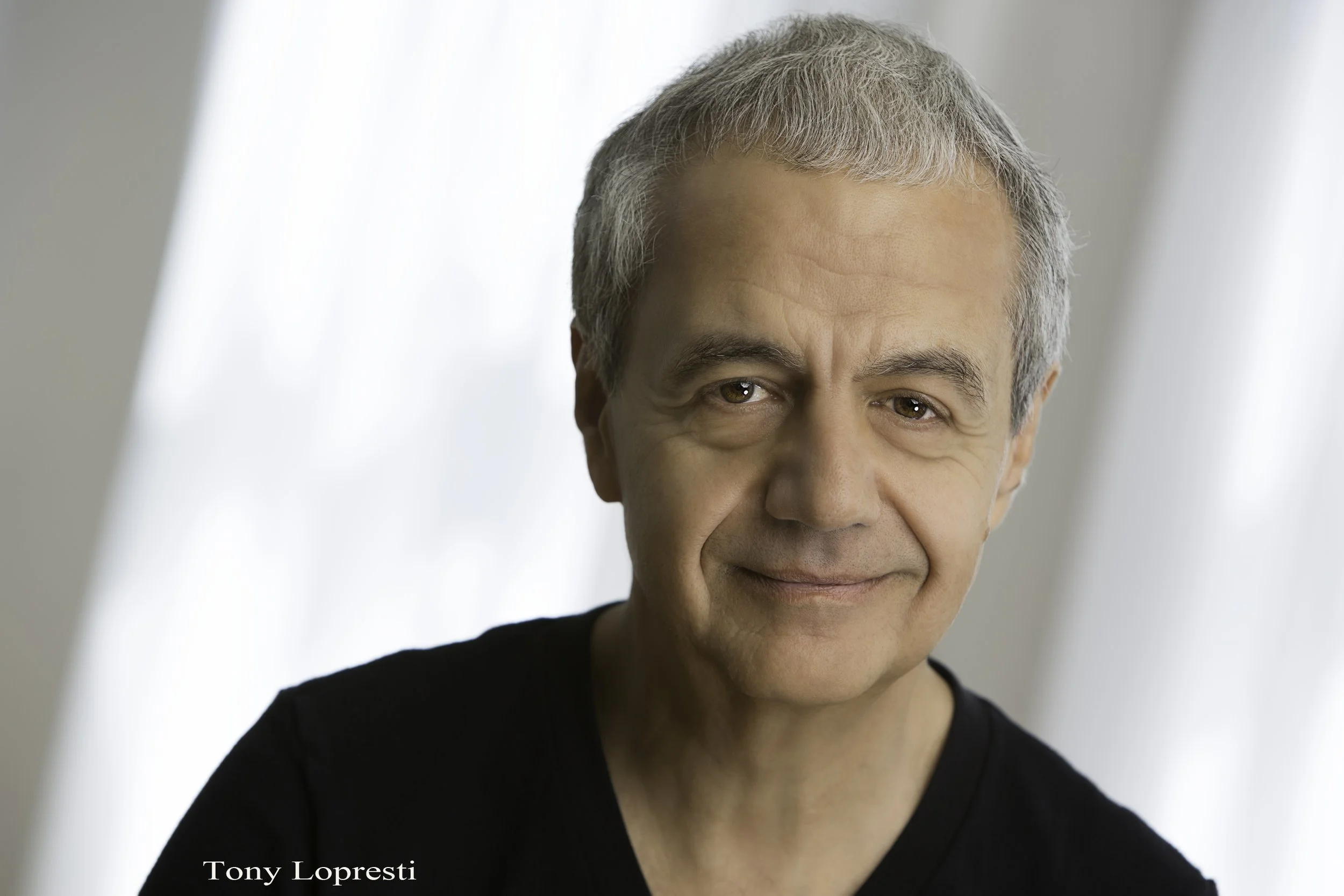 Portrait of an older man with short gray hair and a gentle smile, wearing a black shirt, against a soft-focus light background.