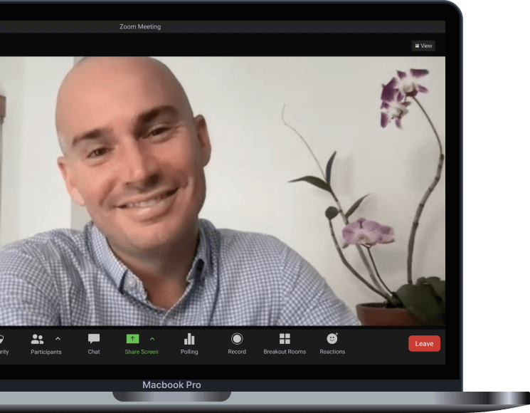 A smiling man in a checkered shirt on a Zoom video call, with a potted orchid plant with pink and purple flowers in the background.