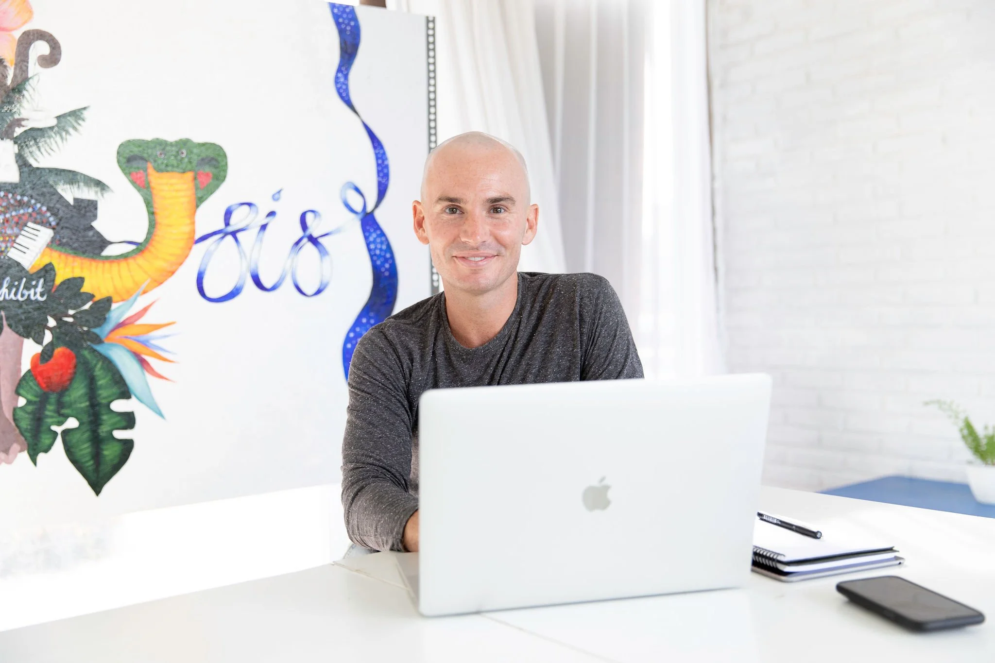 A man sitting at a white desk working on a laptop with a notepad and pen beside him. A colorful mural or art piece is on the wall behind him, and there are white curtains and a white brick wall in the background.