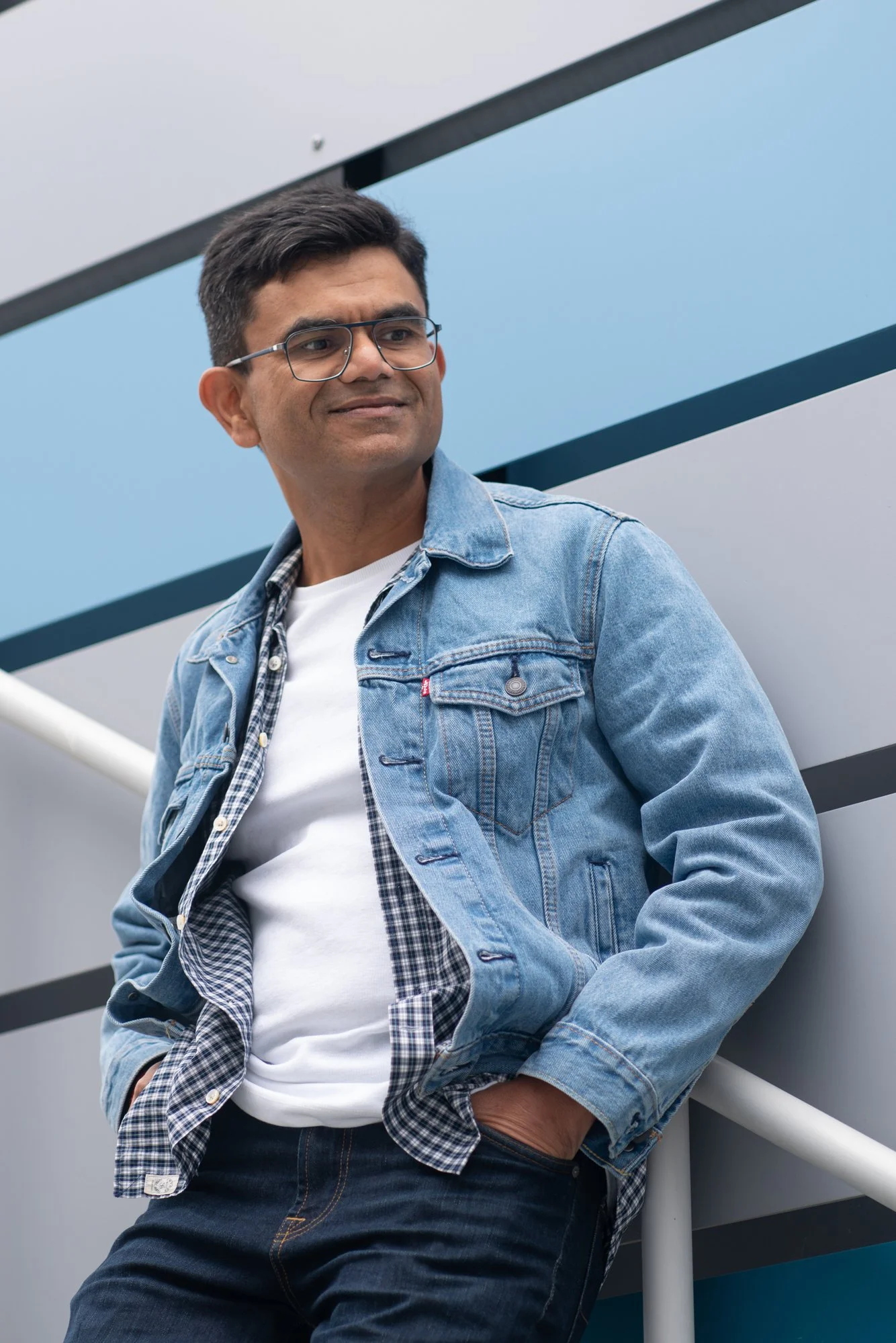 A man wearing glasses, a denim jacket, and a white t-shirt leaning against a railing outdoors.