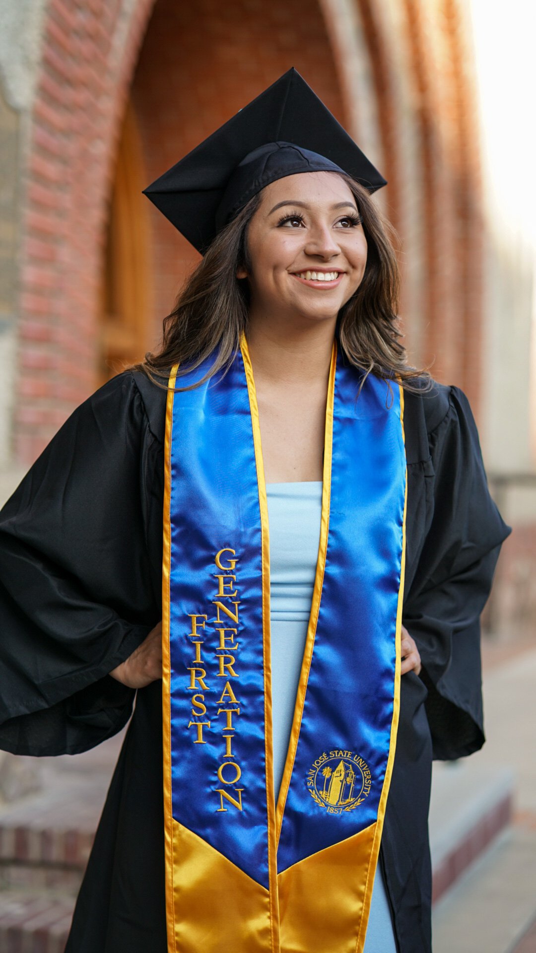 A young woman in a graduation cap and gown with a blue and gold sash that reads 'FIRTS GENERATION' and has a university emblem, standing outdoors.