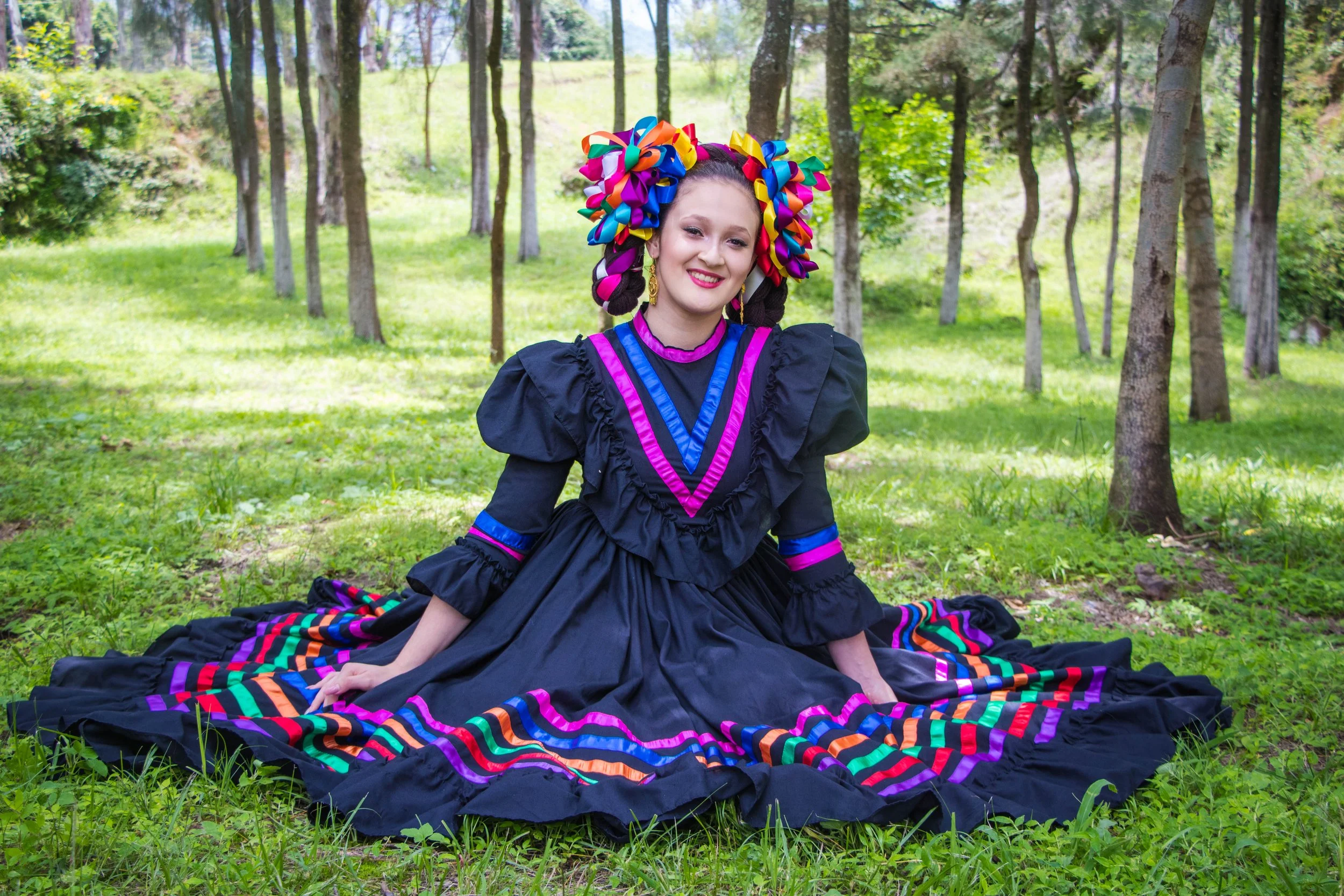 A woman in a traditional Mexican folkloric dress sitting on the grass in a wooded area, with trees and greenery in the background.