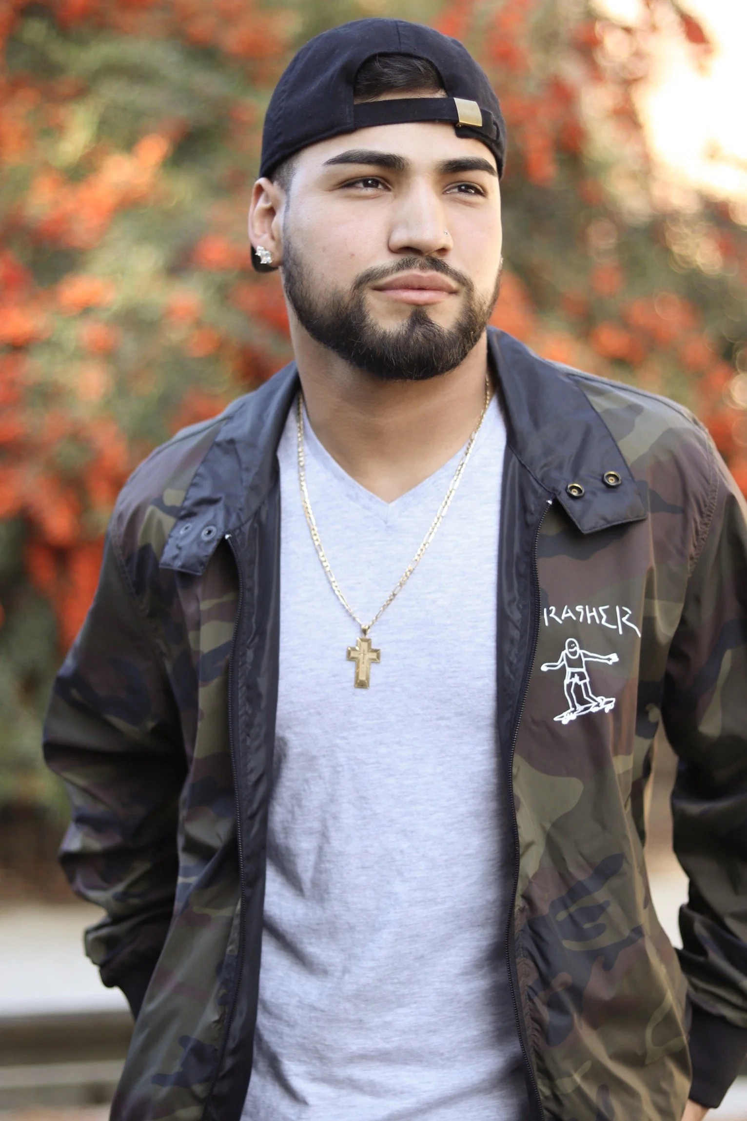 A young man with a beard and earrings, wearing a backwards black cap, a gray T-shirt, and a camouflage jacket with the word 'Thrasher' and a barefoot skateboarder logo, stands outdoors with autumn foliage in the background.