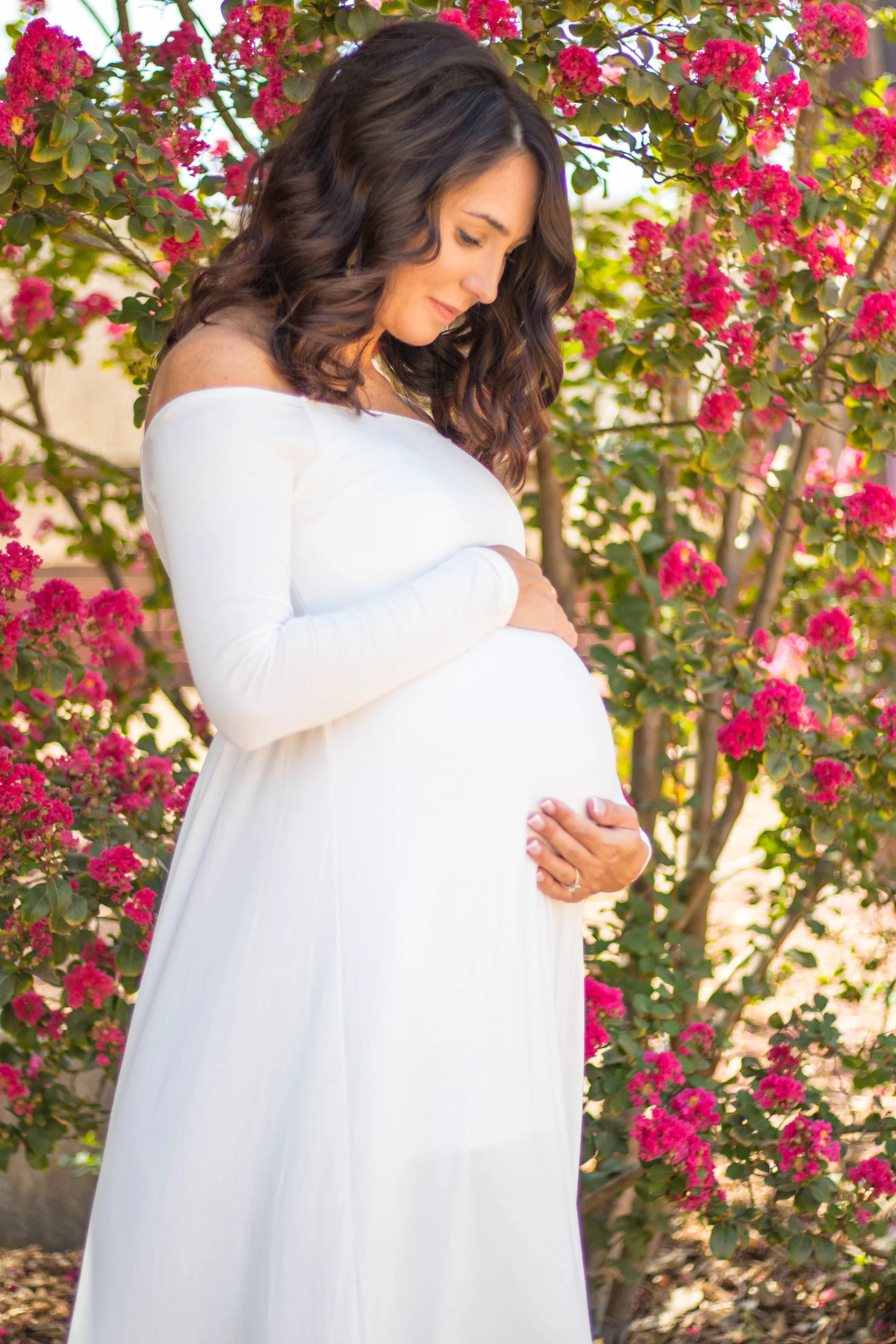 Pregnant woman in a white dress holding her belly, standing outdoors near pink flowering bushes, with her head bowed and eyes closed in a peaceful expression.
