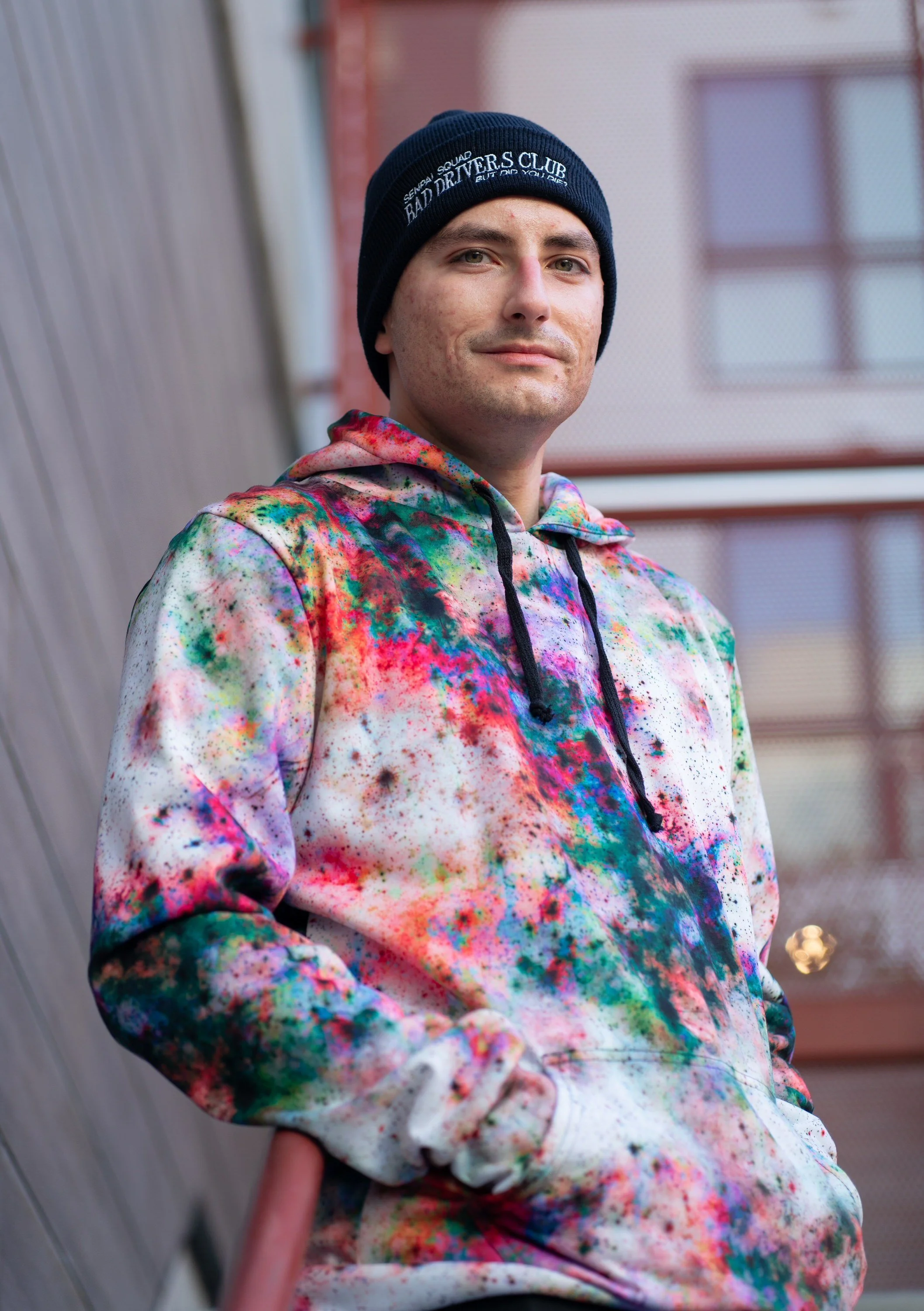 A young man wearing a black beanie and a colorful tie-dye hoodie stands outdoors near a brick building and a wooden wall, looking at the camera with a slight smile.
