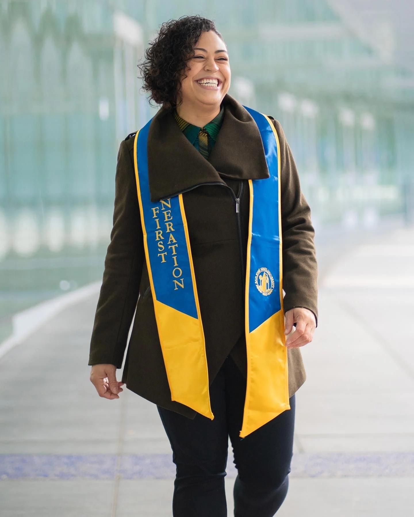 Woman smiling, dressed in dark coat and yellow and blue 'First Generation' stole, walking indoors.
