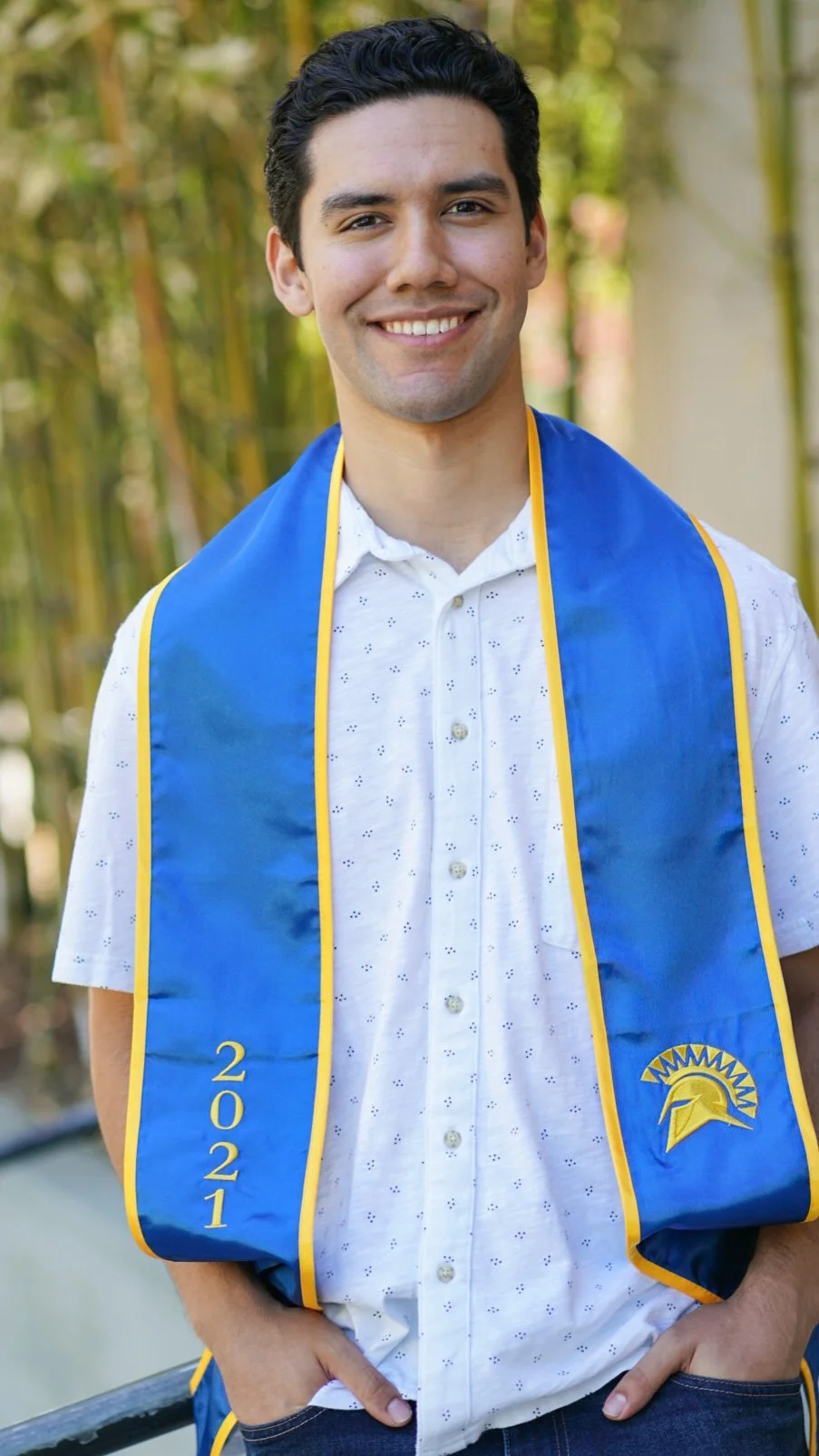Young man wearing a white shirt with small eyelet details and a blue graduation stole with yellow trim and the year 2021. Background of green bamboo stalks.
