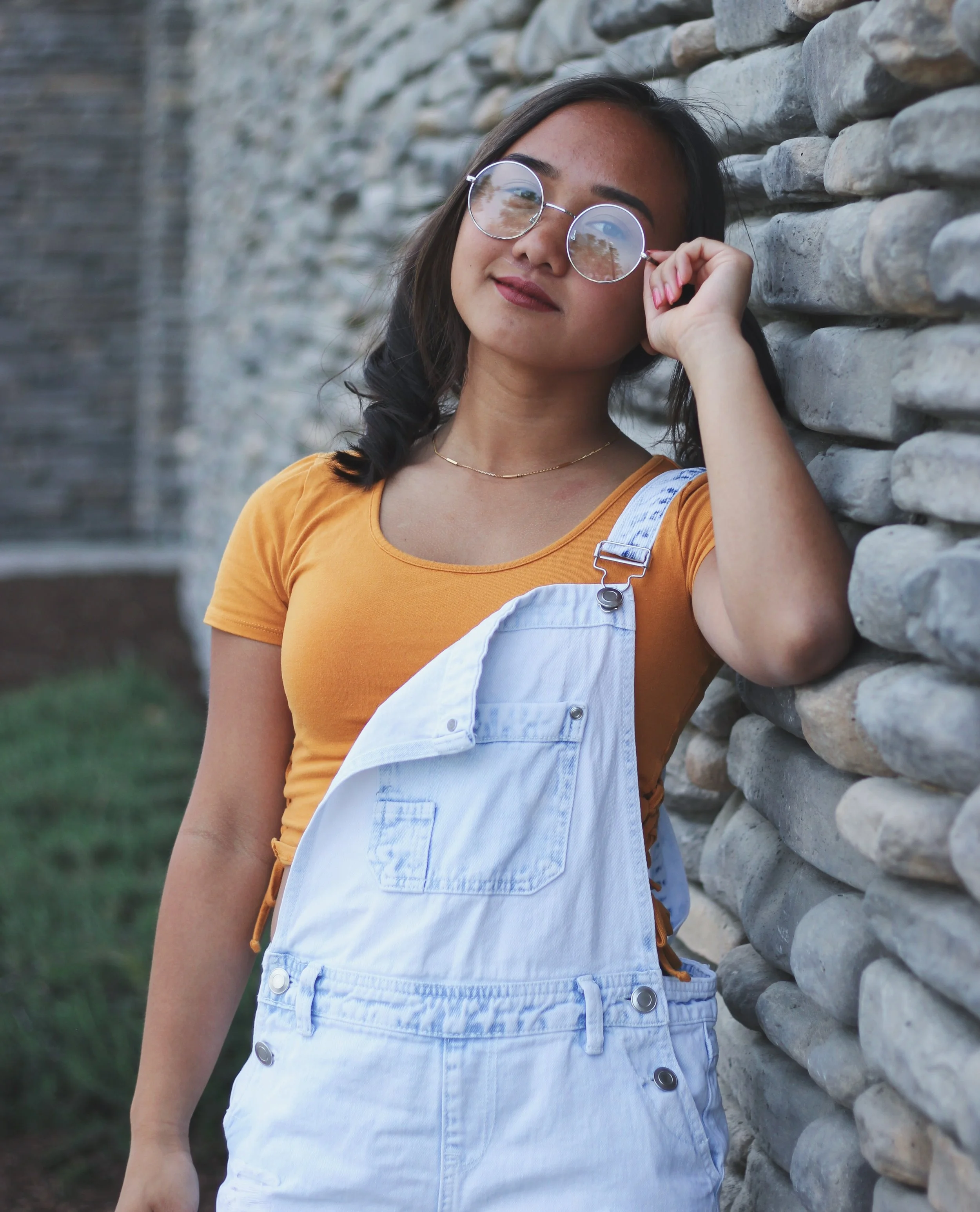 A young woman with glasses, dark hair, and tan skin leaning against a stone wall, wearing an orange shirt and white overalls, looking at the camera.