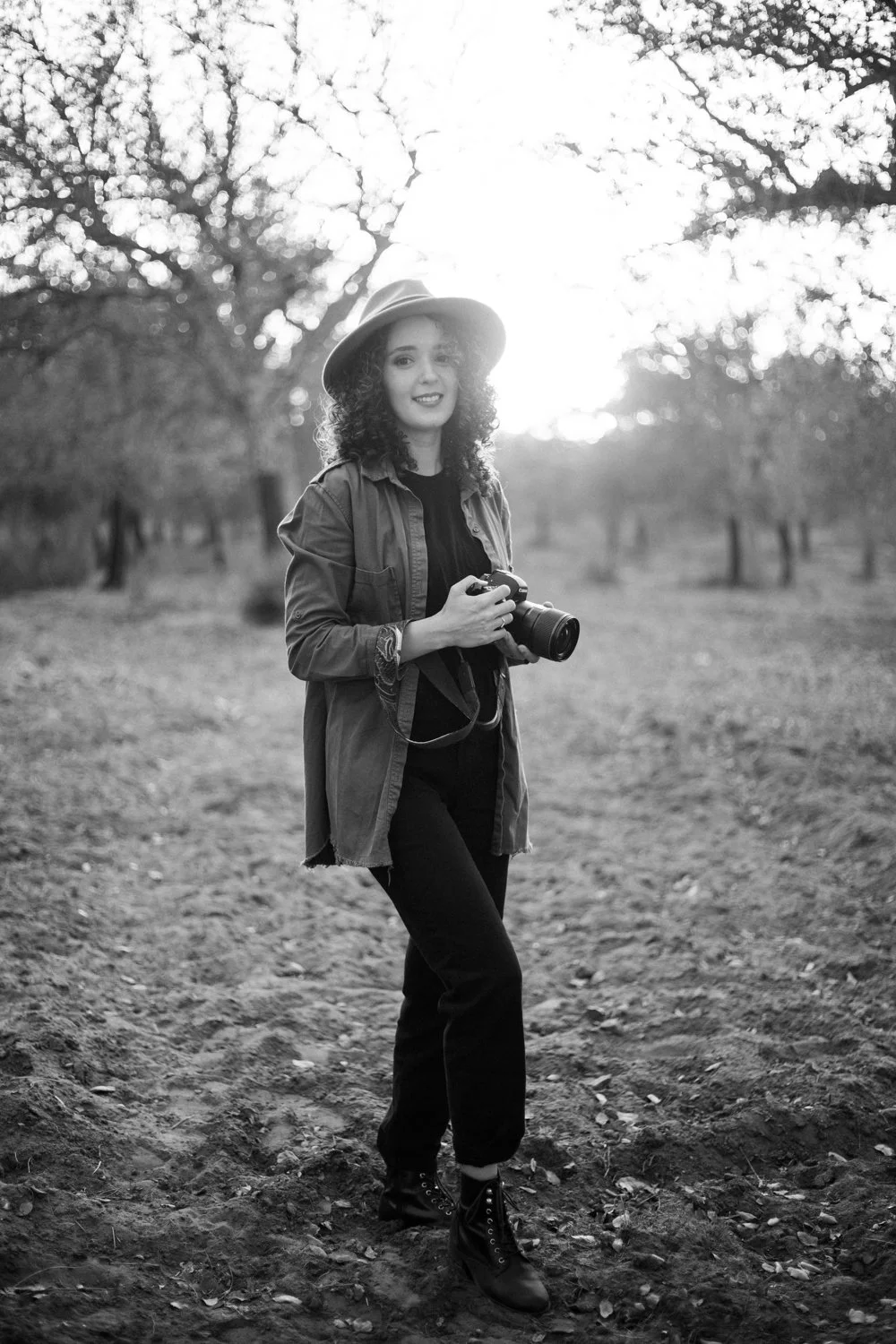 A woman with curly hair wearing a hat, jacket, and boots standing outdoors with trees in the background, holding a camera.