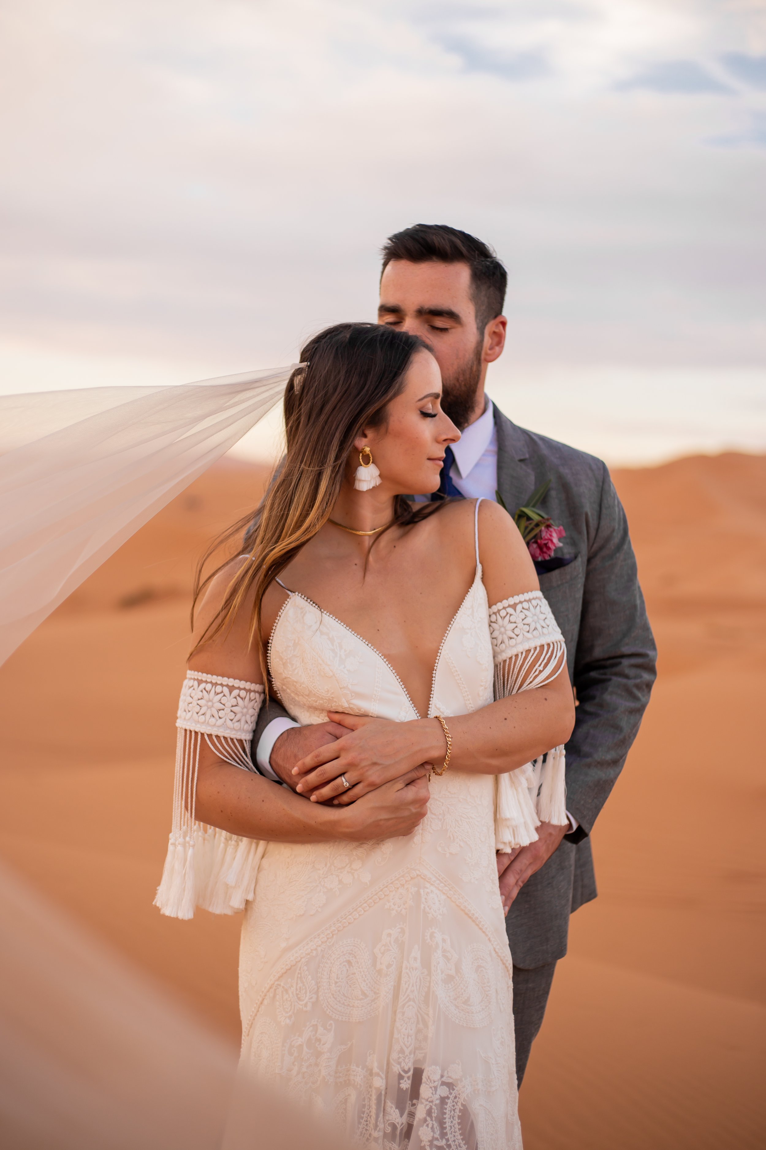 A couple in wedding attire holding each other outdoors with palm trees in the background.
