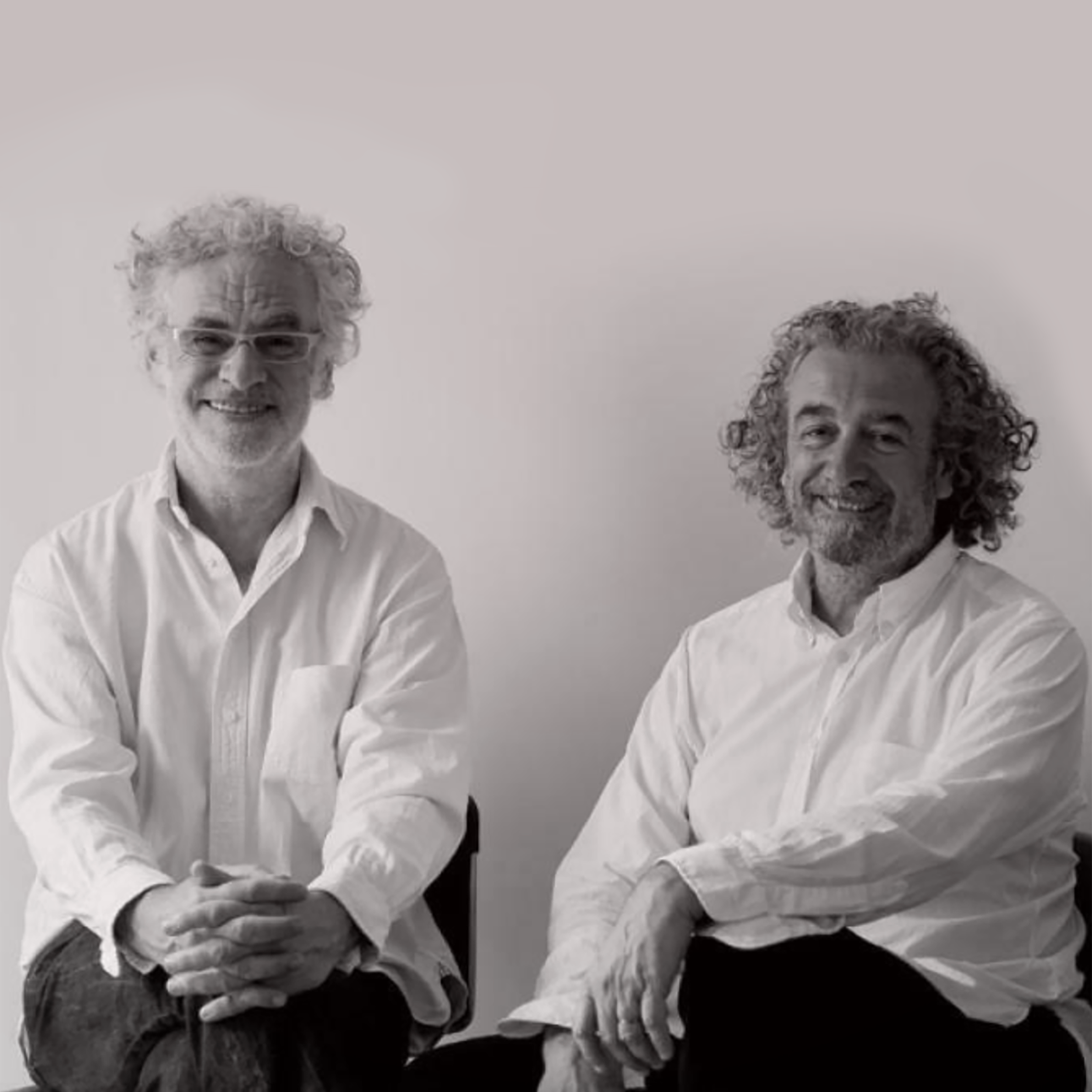Das Bild zeigt zwei Männer, die beide weiße Hemden tragen, sitzend gegen eine helle Wand. Der Mann auf der linken hat lockiges, graues Haar und trägt Brille, während der Mann auf der rechten lockiges, graues Haar und Bart hat.