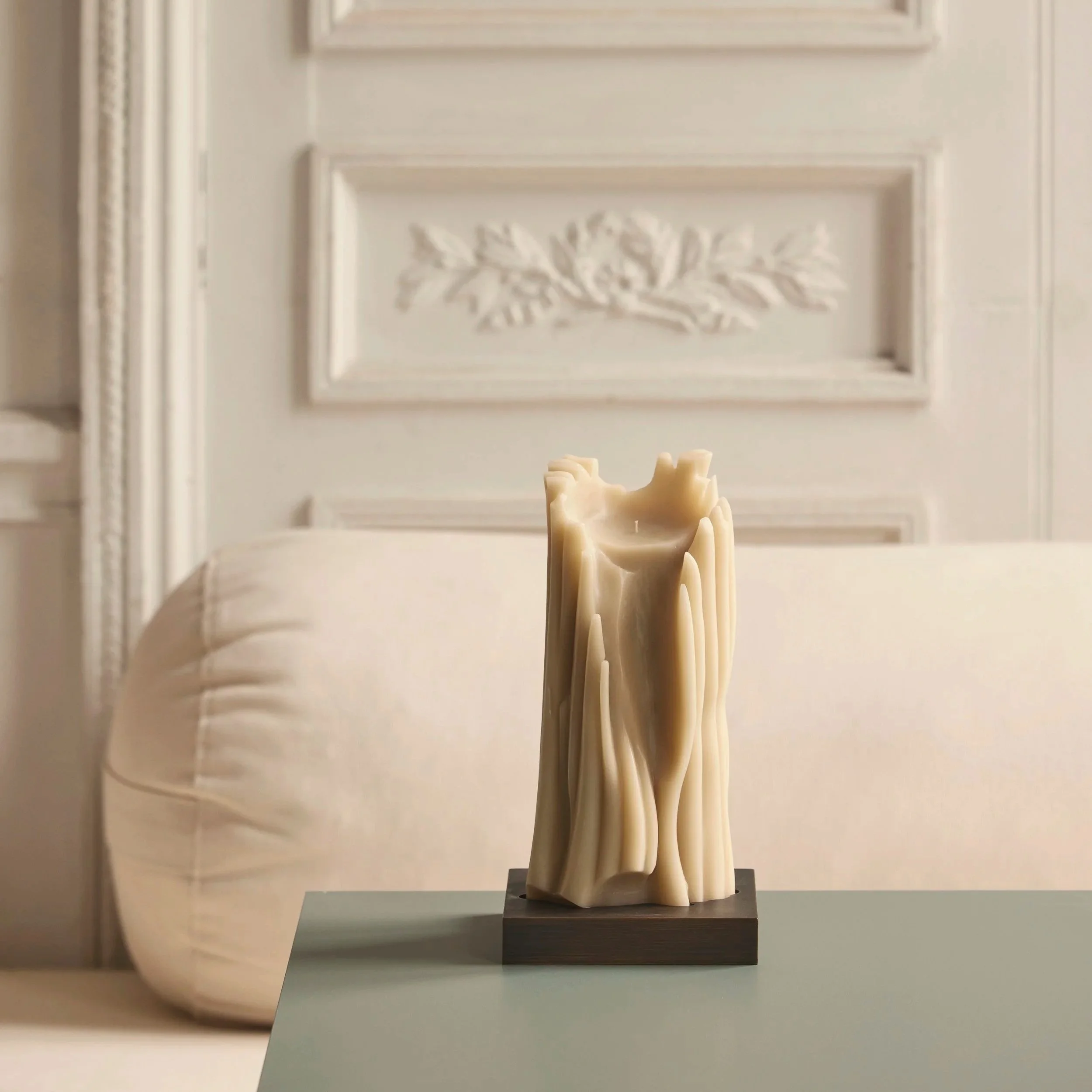 Eine beige Kerze mit einer unregelmäßigen, kunstvollen Oberfläche steht auf einem schwarzen Sockel auf einem Tisch in einem stilvollen Raum mit weißen Wänden und dekorativen Wandpaneelen.