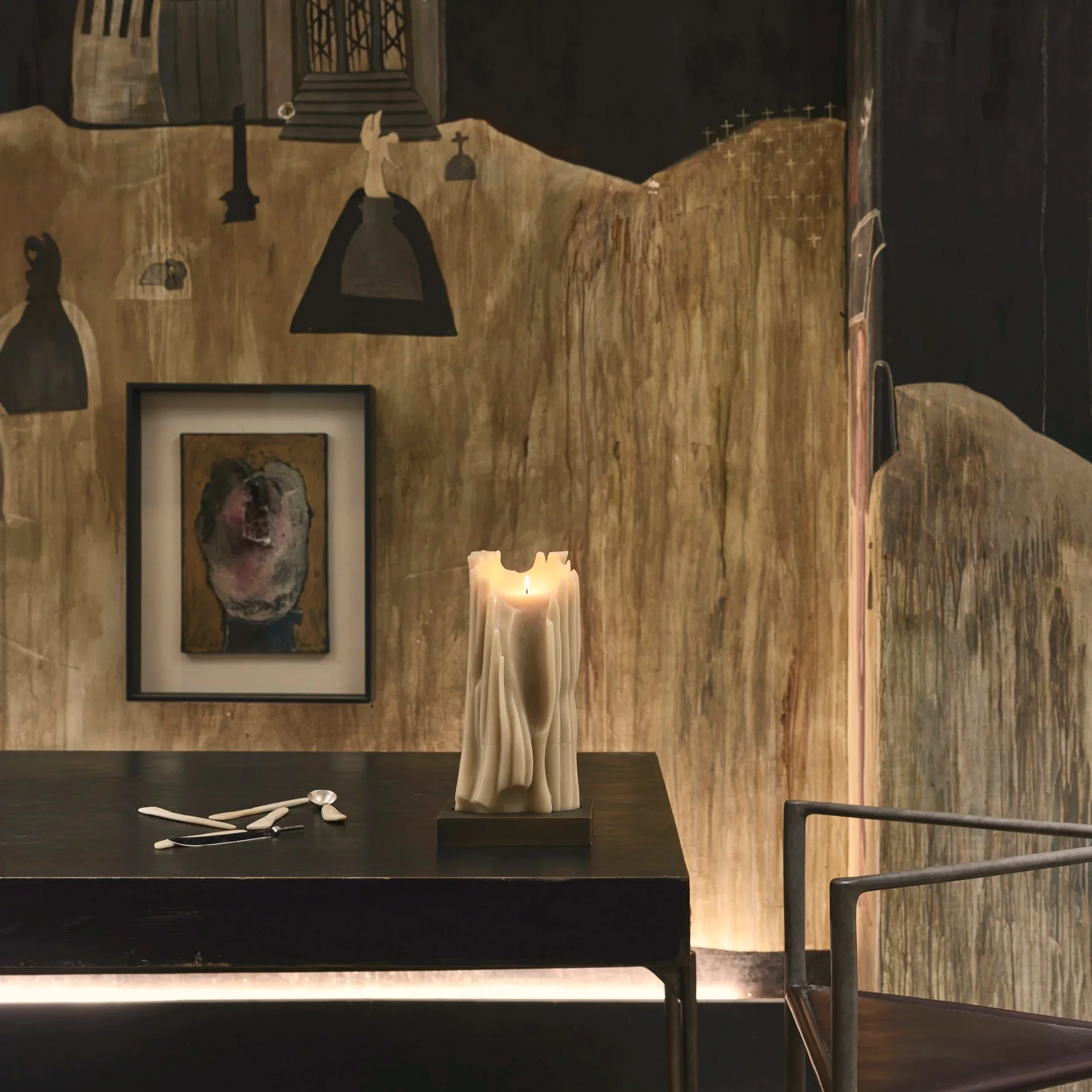 Schwarzer Tisch mit einer brennenden Kerze aus Wachs, weißen Besteckstücken und einem Metallrahmen auf einer Wand mit Kunstwerken im Hintergrund.