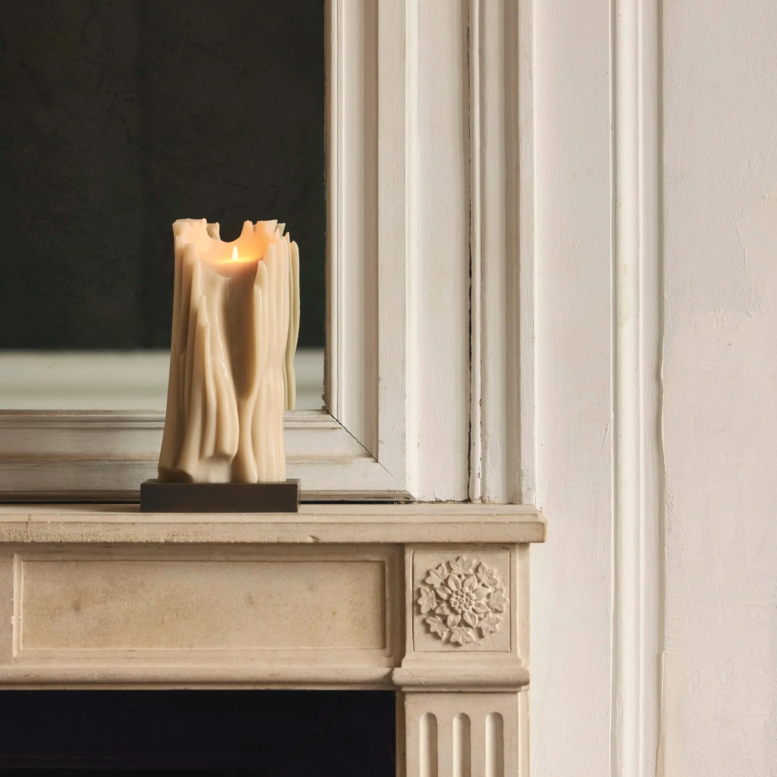 Eine brennende Kerze in Form eines umgestürzten Baumstamms auf einem Kaminsims, davor eine weiße Wand mit verzierter Dekoration.