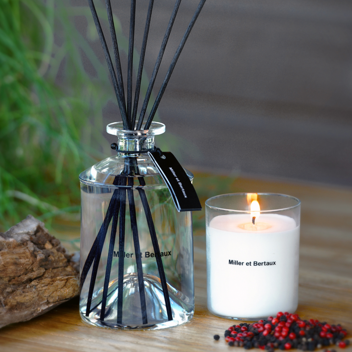 Diffuser mit schwarzen Stäbchen, brennende weiße Kerze in einem Glas mit der Aufschrift 'Miller et Bertaux', neben einem Stück Holz und Pfefferkörner