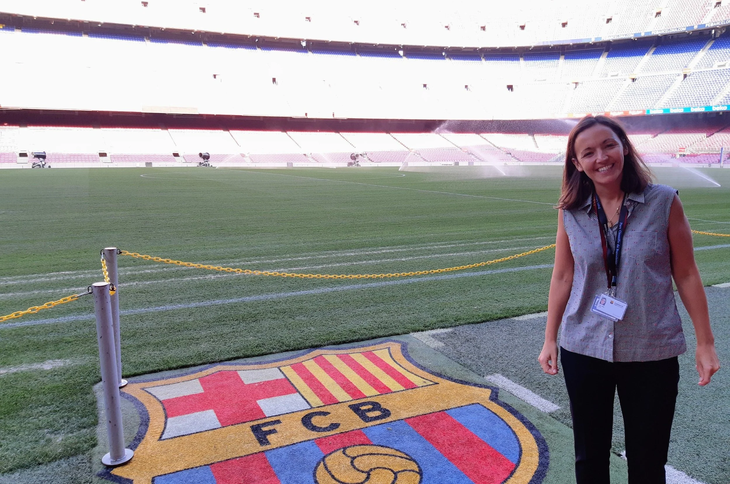 Lucy Mills, founder of Ready Sport Global, smiling at the camera standing on FC Barcelona's Camp Nou stadium with FC Barcelona's logo rug beneath her feet. The background shows stadium seats and spray water from sprinklers on the field.