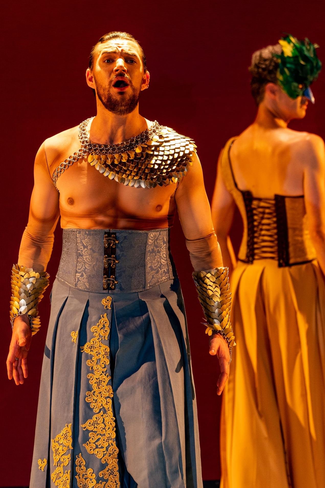 A man with long hair and a beard singing on stage, wearing a costume with metallic shoulder pieces, arm cuffs, a corset, and pants with gold embroidery, next to a woman in a yellow outfit with a floral headpiece.