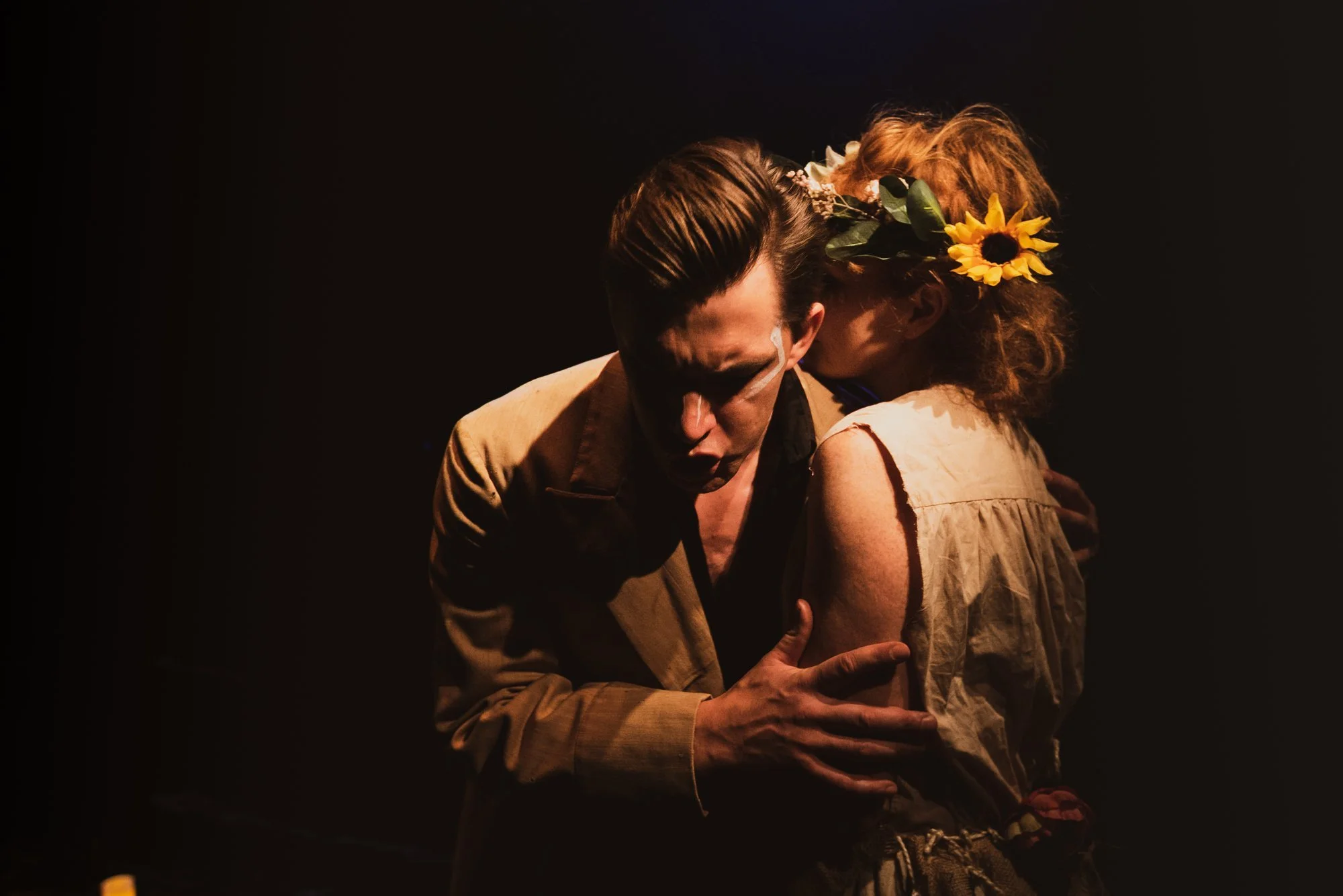 A theatrical performance showing a man and a woman in an emotional embrace, with the woman whispering into the man's ear. The woman has a sunflower in her hair.