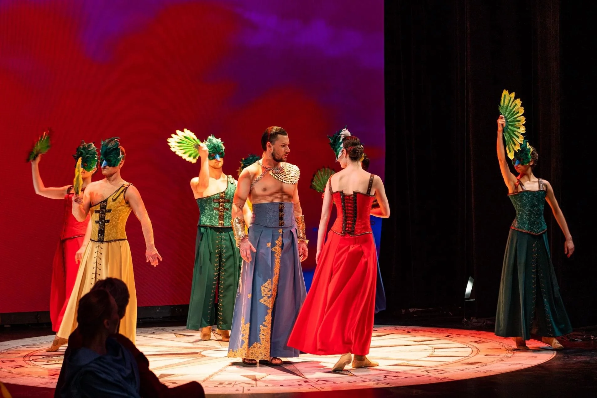 Performers in colorful costumes and feather masks on stage during a theatrical performance.