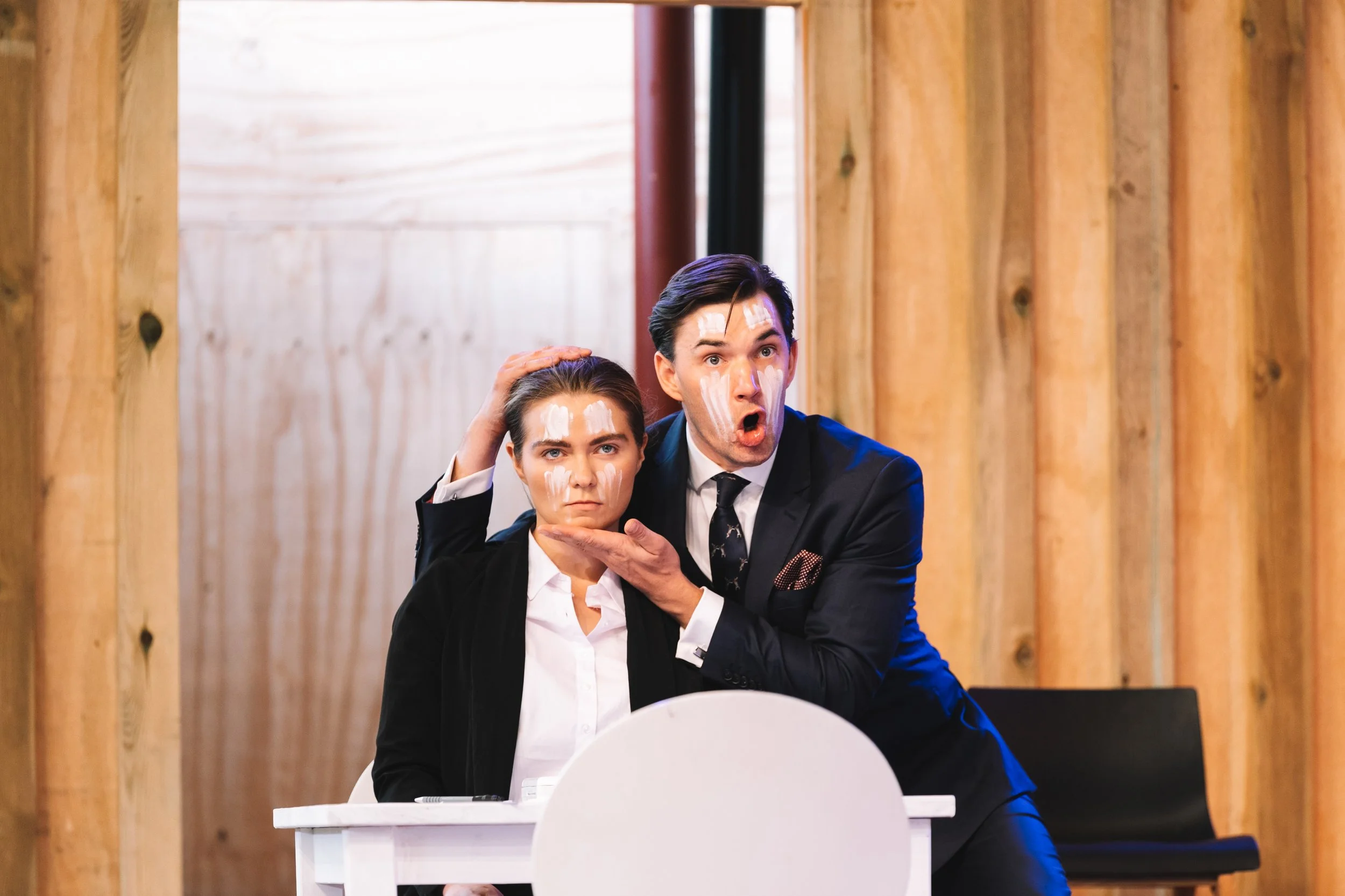 A man in a black suit and a woman in a white shirt with a black blazer sit at a white desk with exaggerated expressions on their faces. The man appears surprised or shocked, with his mouth open and eyebrows raised, while the woman looks serious or an