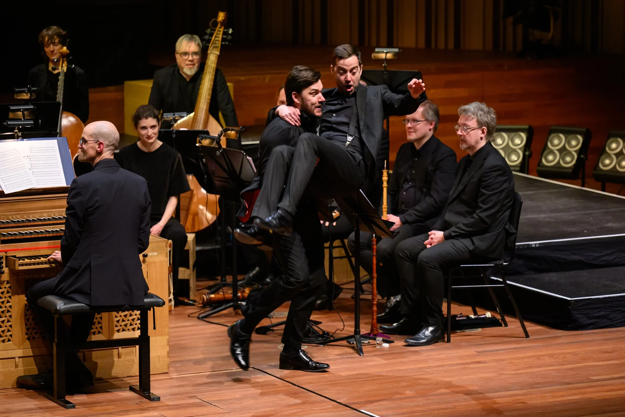 Orchestra members on stage with two men in suits at center, one lifting the other in celebration during a concert.
