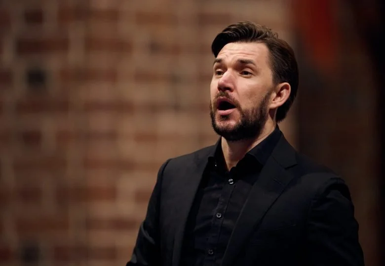 A man with dark hair and a beard, wearing a black suit and black shirt, appears to be singing or speaking passionately in front of a brick wall.