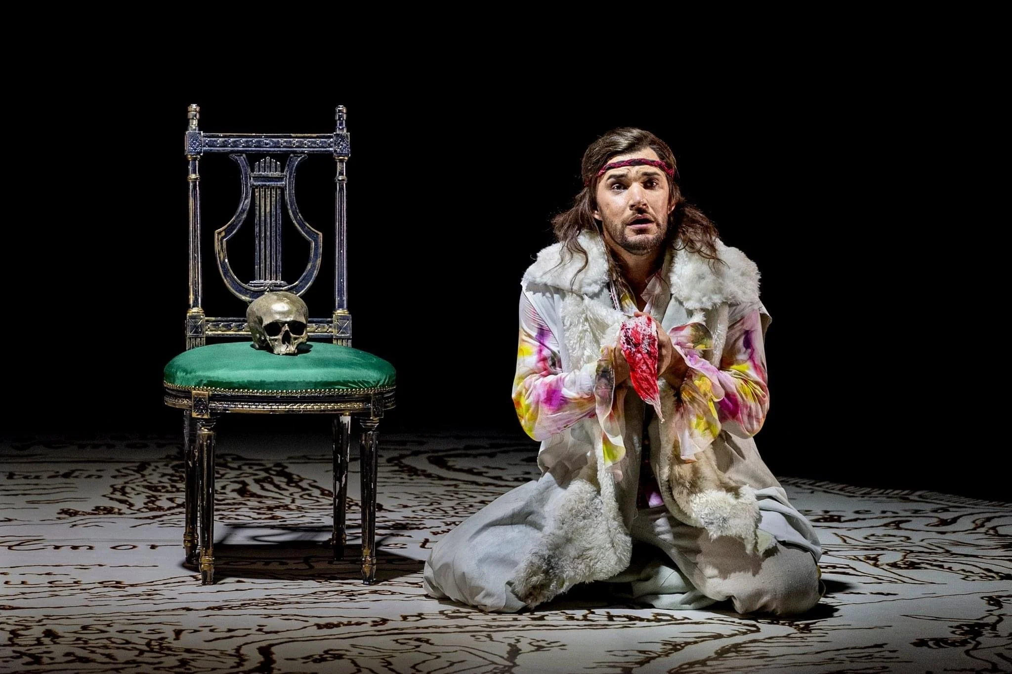 A man dressed in colorful, bohemian clothing kneeling on a patterned floor, holding a red cloth, with a green-velvet cushioned chair behind him, on which a skull rests. The background is black.
