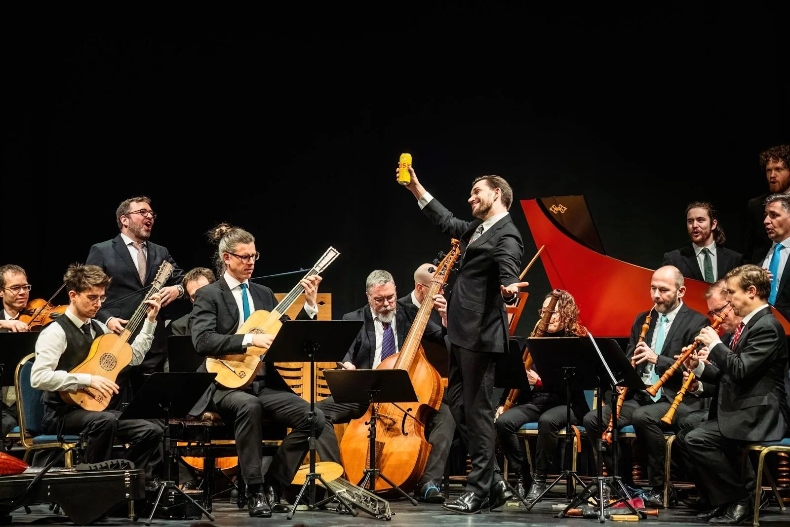 Orchestra performing on stage with a conductor holding a yellow water bottle, surrounded by musicians playing various string and wind instruments.