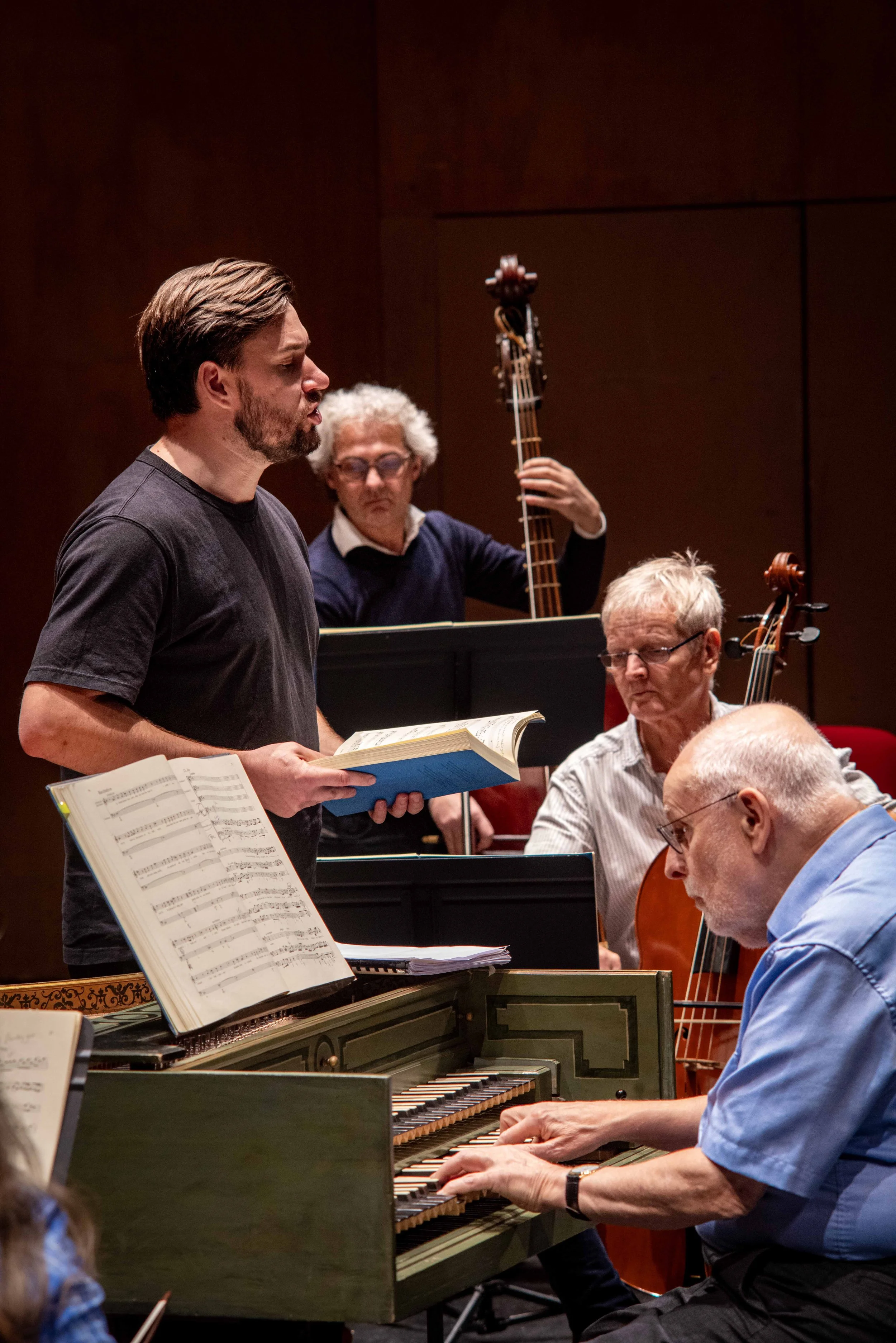 Group of musicians practicing in a room, with a man standing and singing, playing sheet music, and three others playing piano, violin, and double bass.
