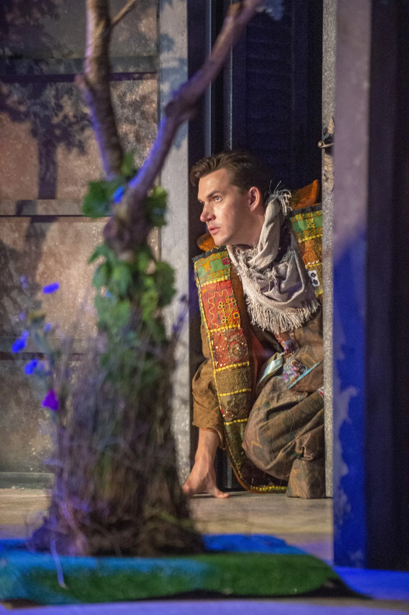 A man with slicked-back hair and light skin is looking to the side while crouching on the ground, wearing a colorful patchwork jacket and a scarf. He is beside a wall, with a tree and some purple flowers in the foreground and a dark, decorated enviro