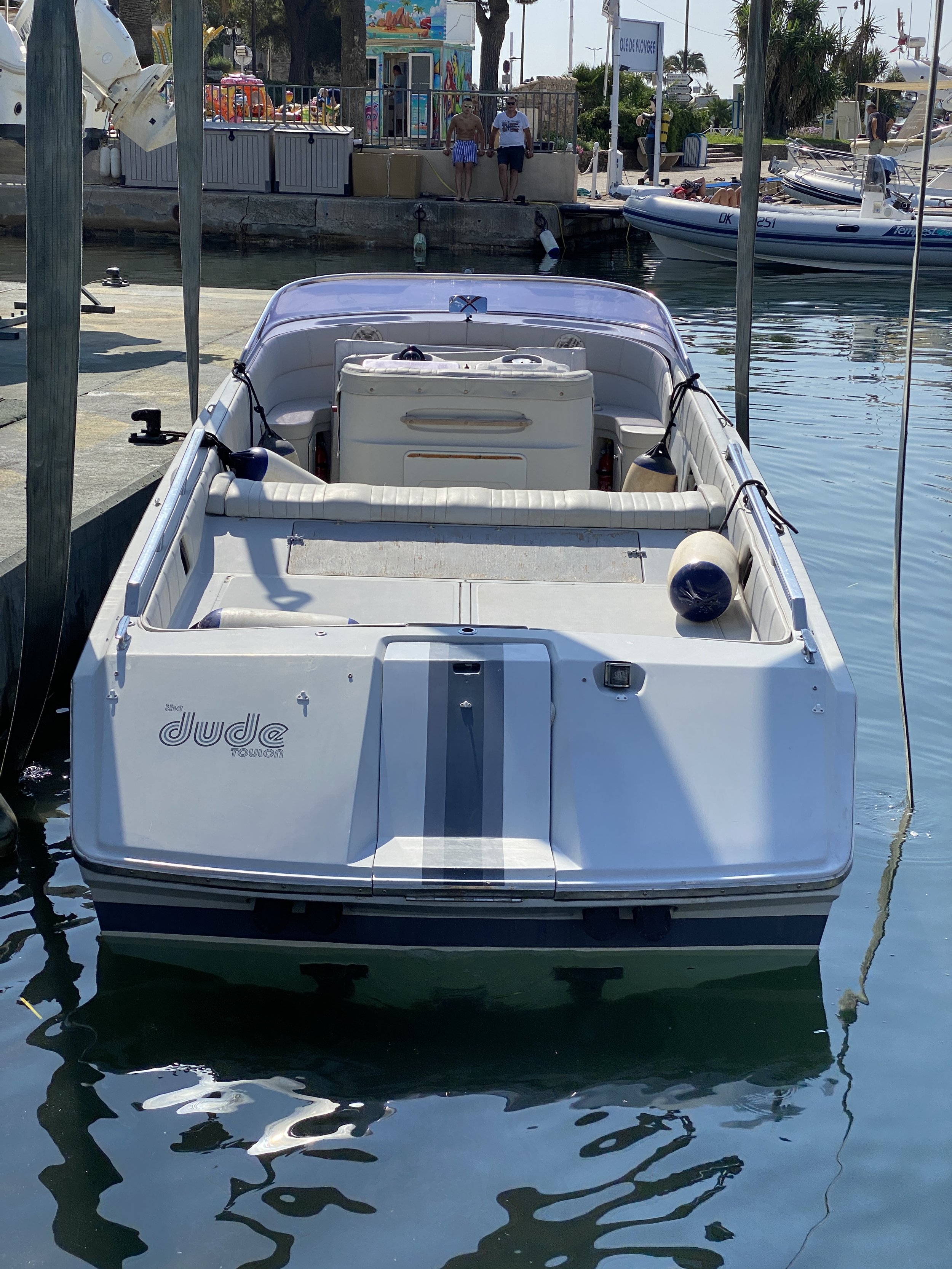 A white boat named 'The Jude' is docked at a marina with a few people standing on the walkway in the background, and other boats floating in the water nearby.