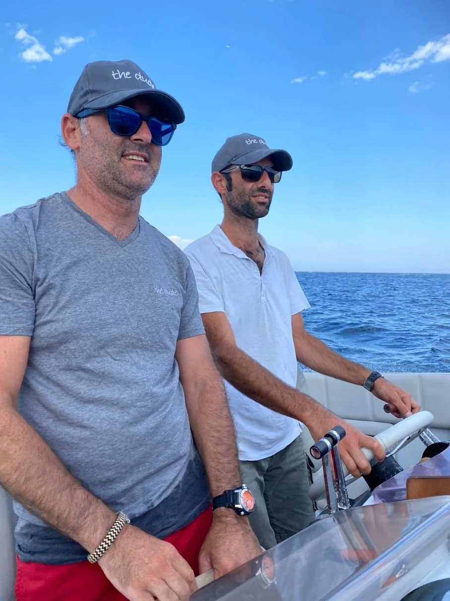 Two men wearing sunglasses and hats on a boat steering in the ocean under a blue sky.