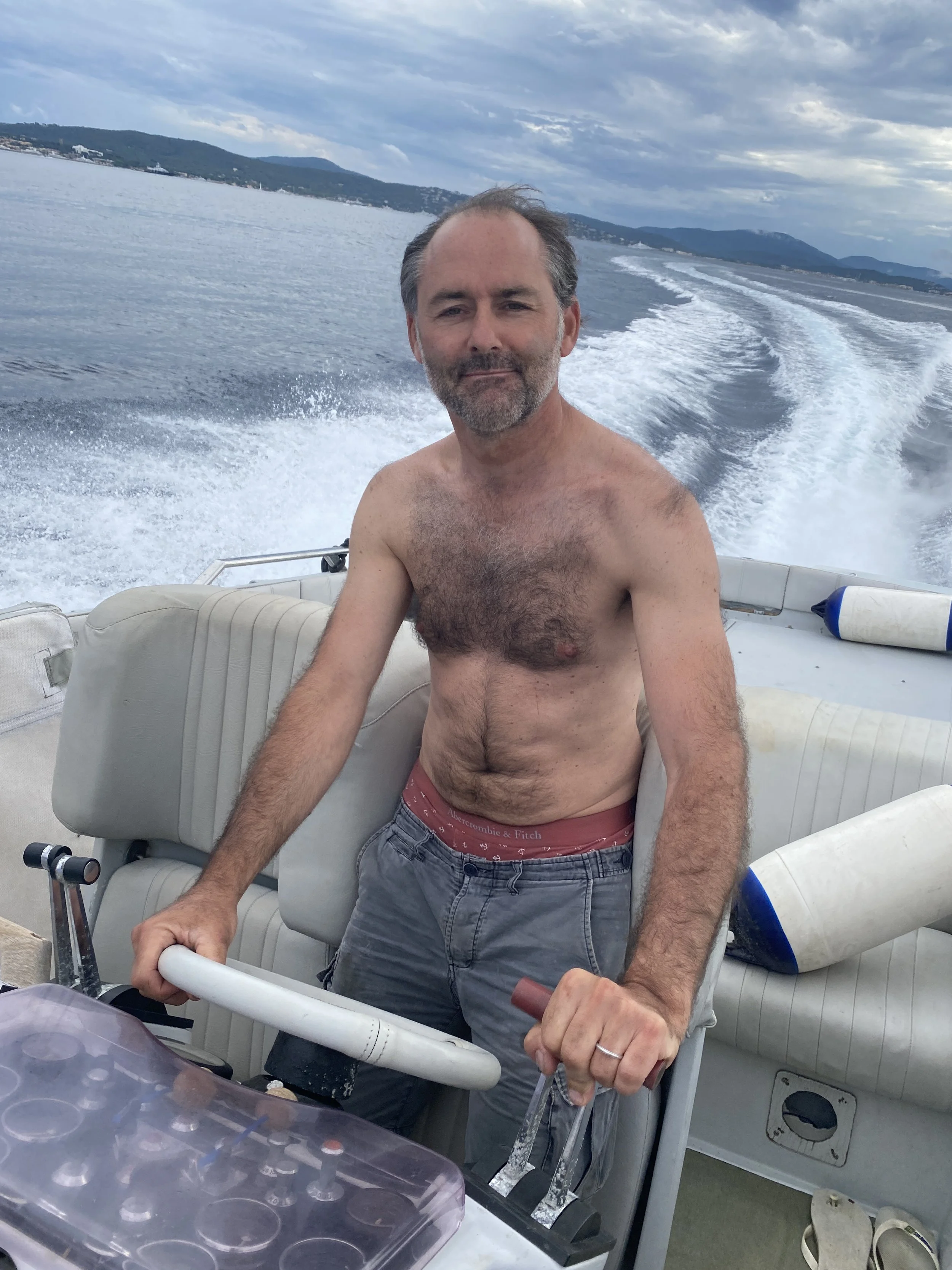 A shirtless man with gray hair and a beard stands at the helm of a boat on a body of water, with hills and cloudy sky in the background.