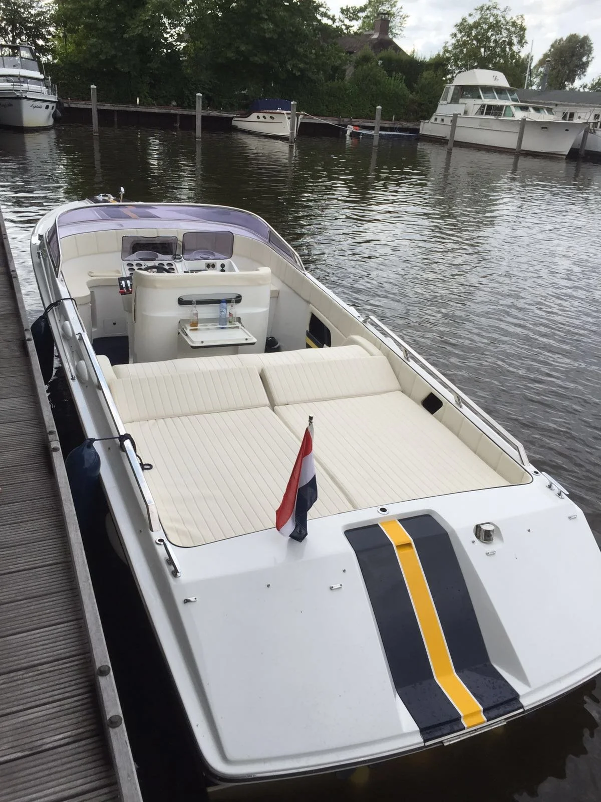 A white motorboat with a black and yellow stripe on the hood and a Dutch flag at the front, docked at a wooden pier on a calm waterway. Several other boats are moored in the background, with trees and houses along the shoreline.