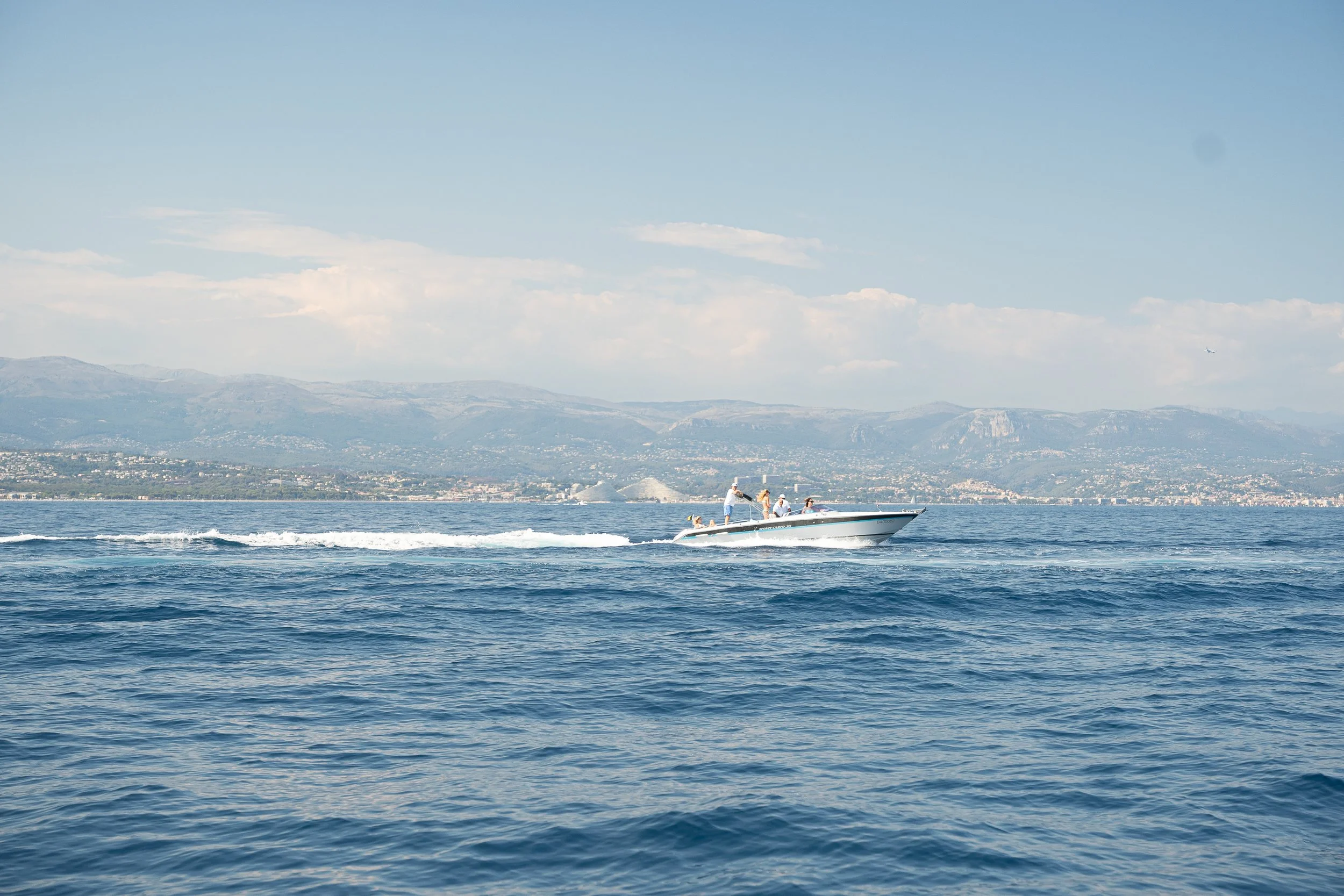 A speedboat with four people on board speeding across a calm blue ocean with a distant shoreline and mountains in the background.