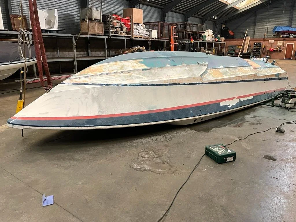An indoor storage or workshop space with a large, upside-down boat resting on the floor. The boat appears to be in the process of restoration or repair, with visible patches of sanding and primer. Various shelves and storage bins are visible in the b