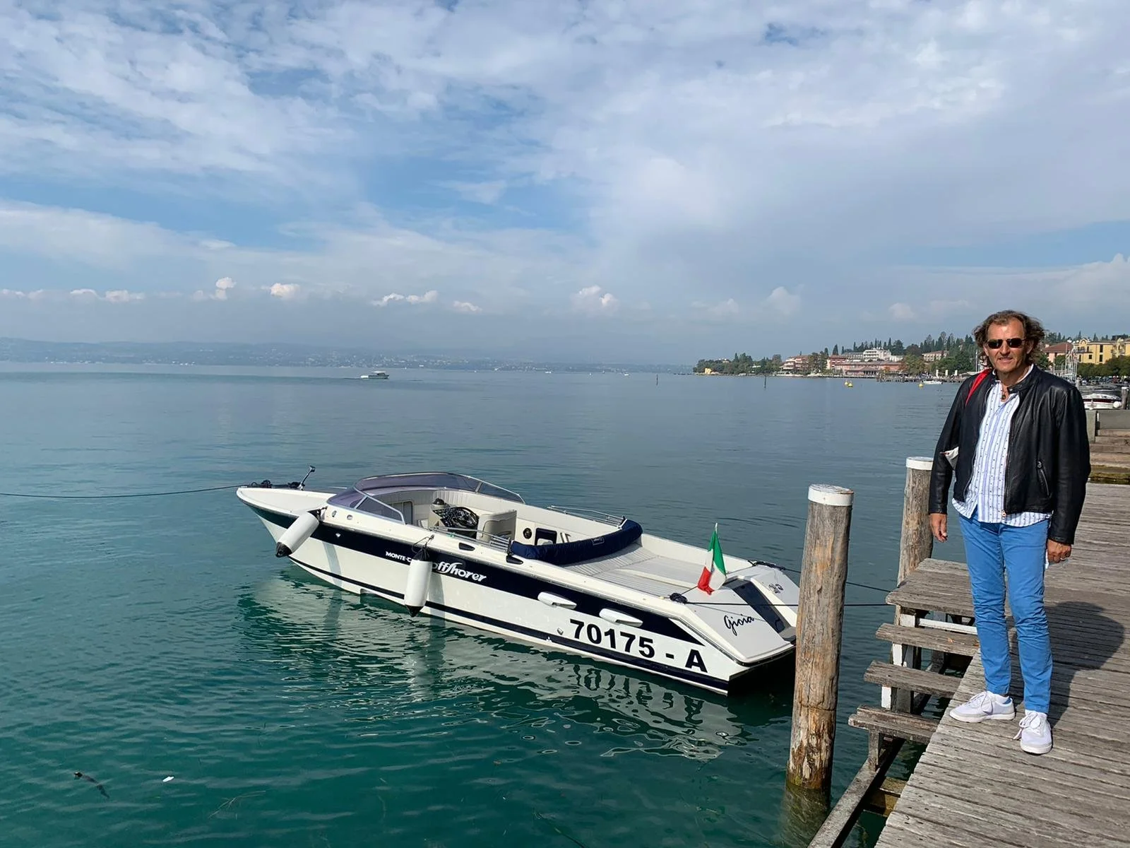 A man in a leather jacket and sunglasses stands on a wooden dock next to a boat in a calm lake with a distant shoreline and cloudy sky.