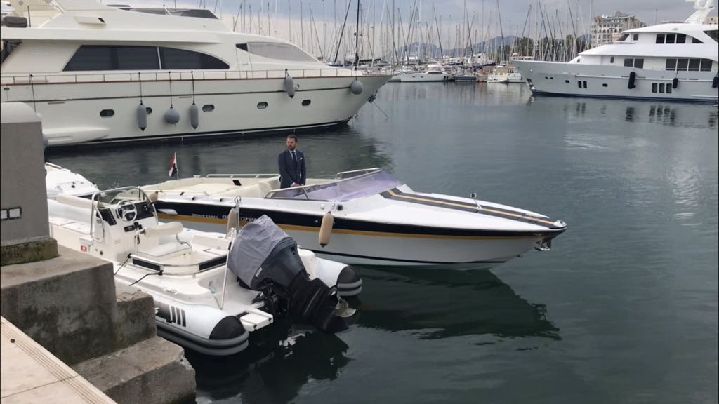 A man in a suit standing on a dock near a small white and black speedboat with an outboard motor, overlooking a marina with large yachts and sailboats.