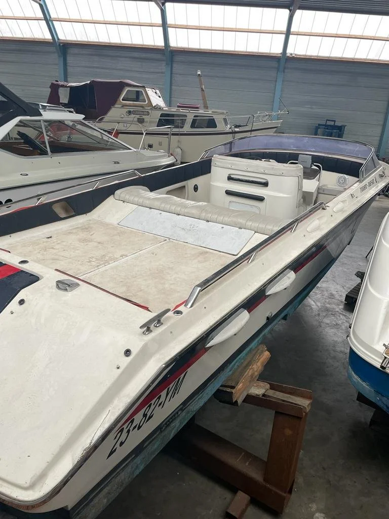 A small white motorboat with a black stripe and red accents inside a storage facility. The boat has seating and is supported by wooden blocks. Other boats are visible in the background.