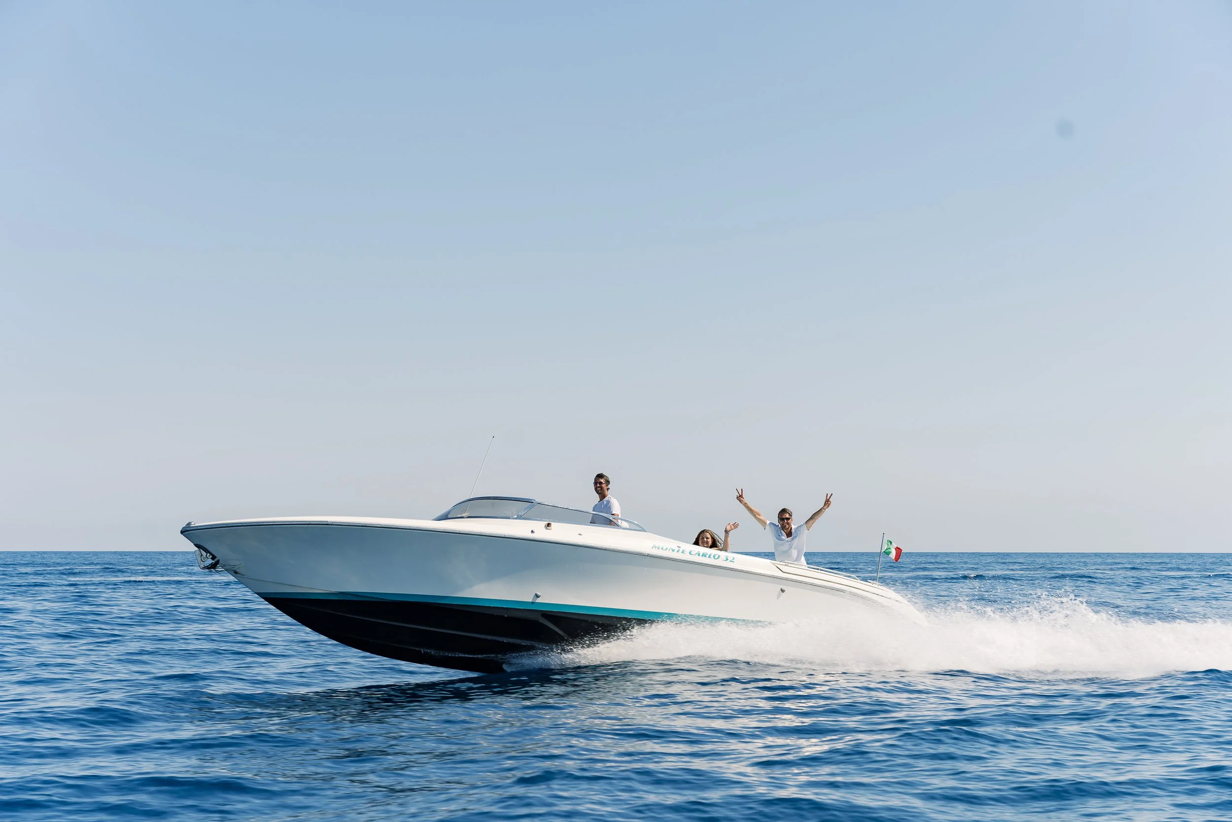 Three people on a white motorboat speeding across the water, with one person raising their arms in joy and another making a peace sign, under a clear blue sky.