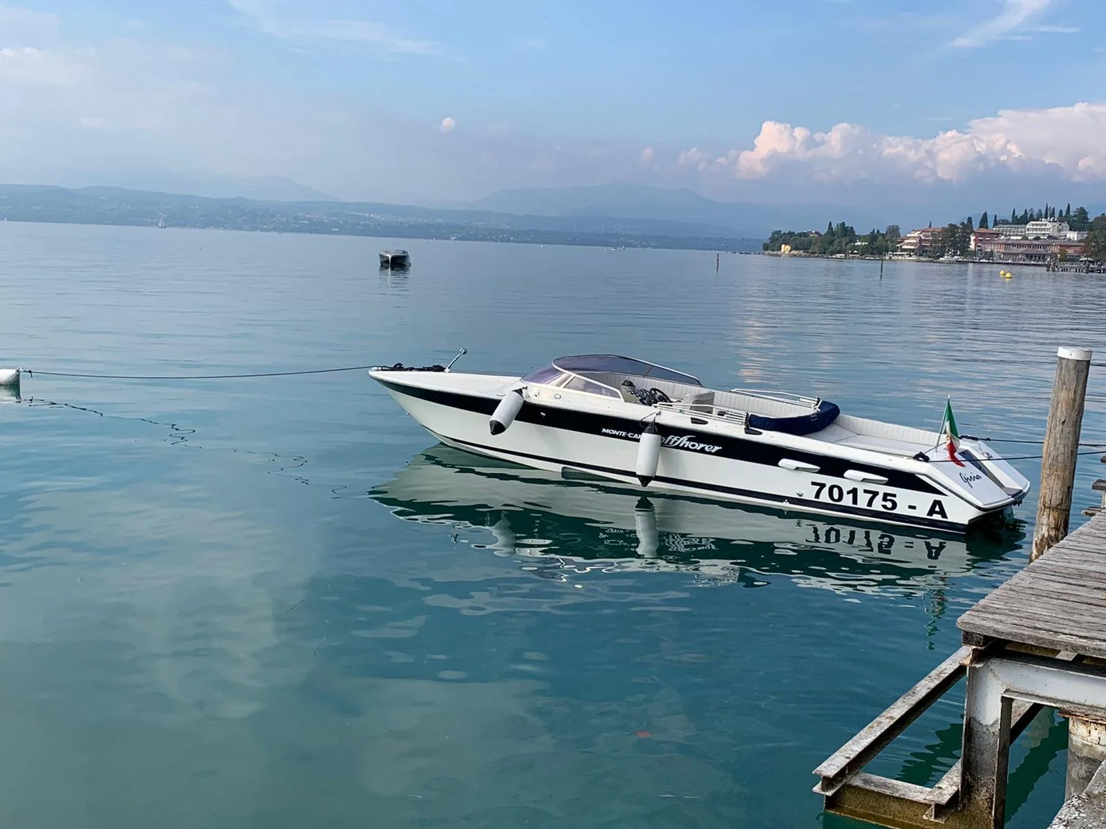 A white motorboat with black accents and the registration number 70175-A is moored to a wooden dock on a calm lake. The background features distant mountains, a partly cloudy sky, and a lakeside town.