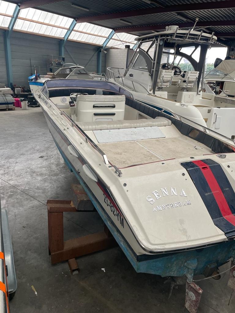 A white motorboat with blue and red stripes inside a large boat storage facility, supported by a wooden stand, with other boats in the background.