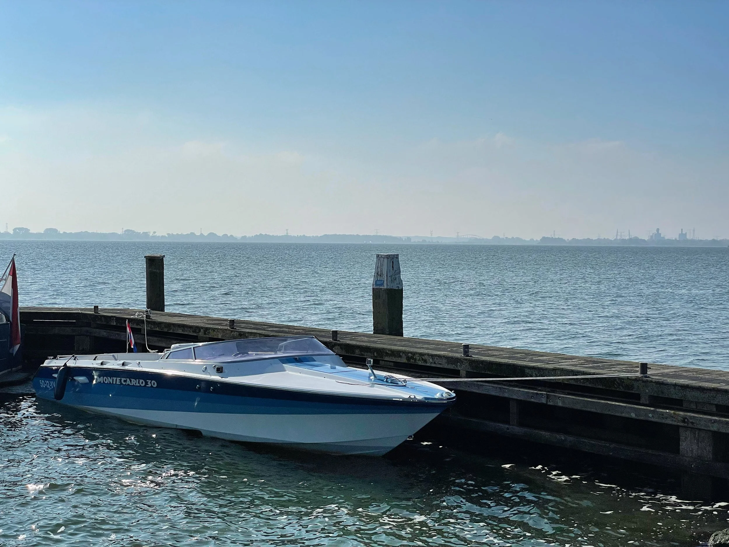 A white and blue speedboat docked at a wooden pier with calm water and a distant shoreline under a clear blue sky.