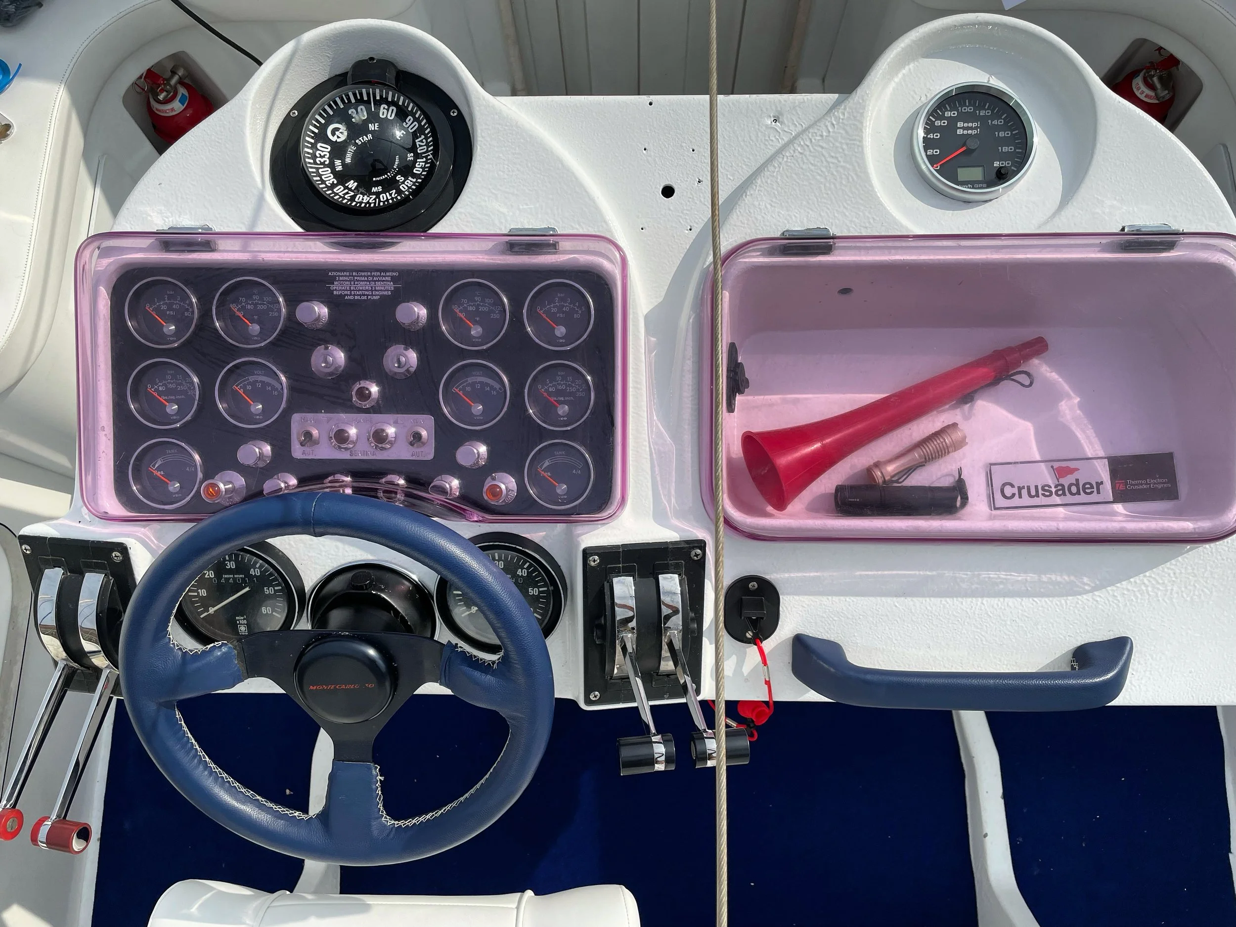 Boat dashboard with gauges, steering wheel, and a clear plastic box containing a red horn, flashlight, and other items, with a Crusader sticker.