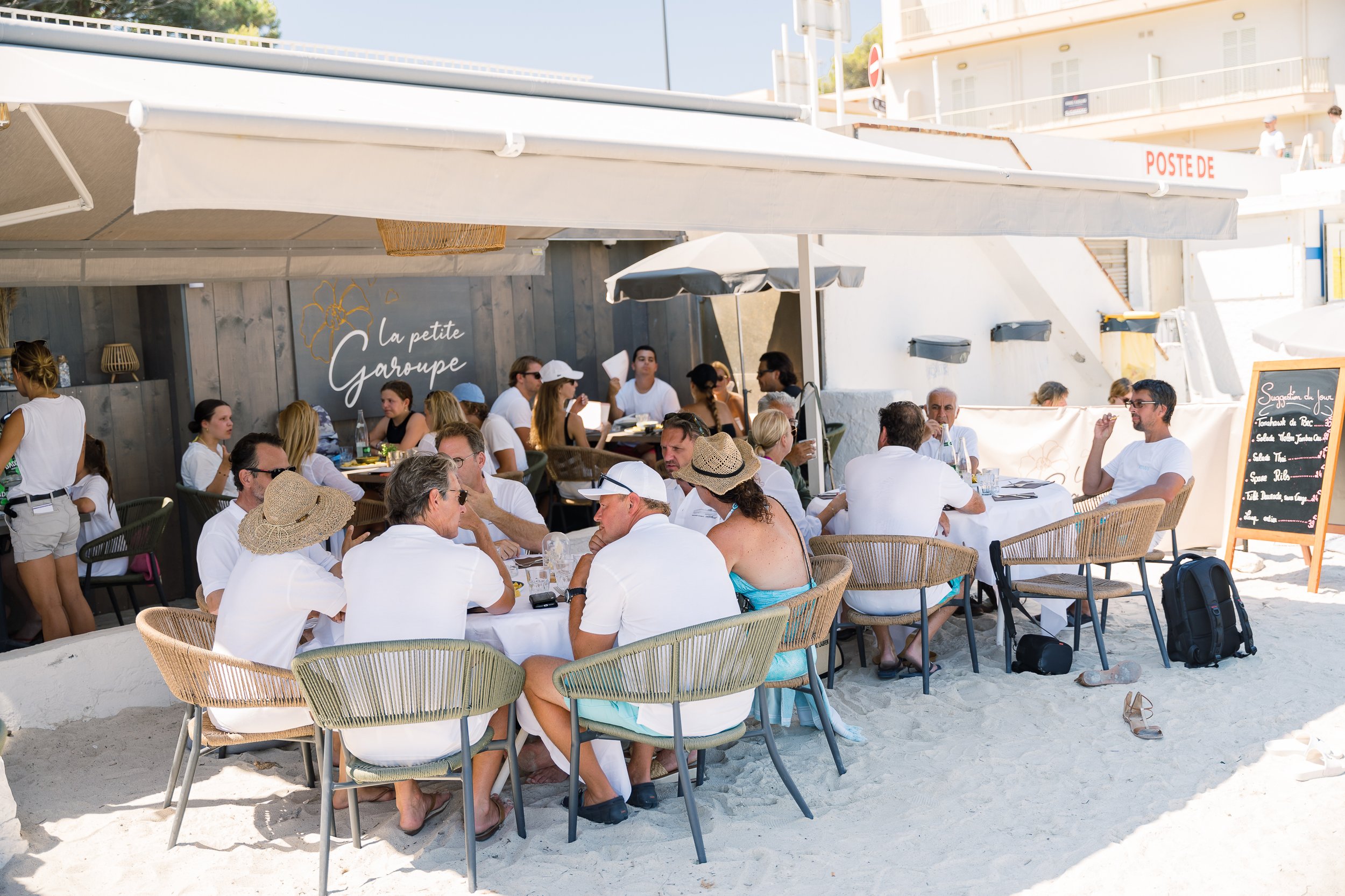 People dining outdoors at a beachside restaurant, some wearing hats and sunglasses, with sandy ground and a white building in the background.