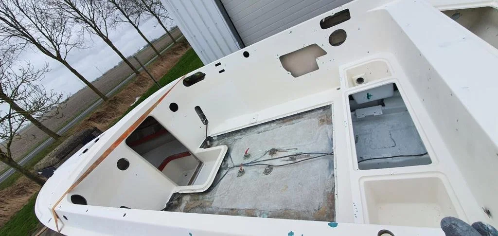 The image shows the interior of a boat that is under repair or renovation, with a mostly empty and clean deck, exposed wires, and open compartments, situated outdoors near some trees and a building.