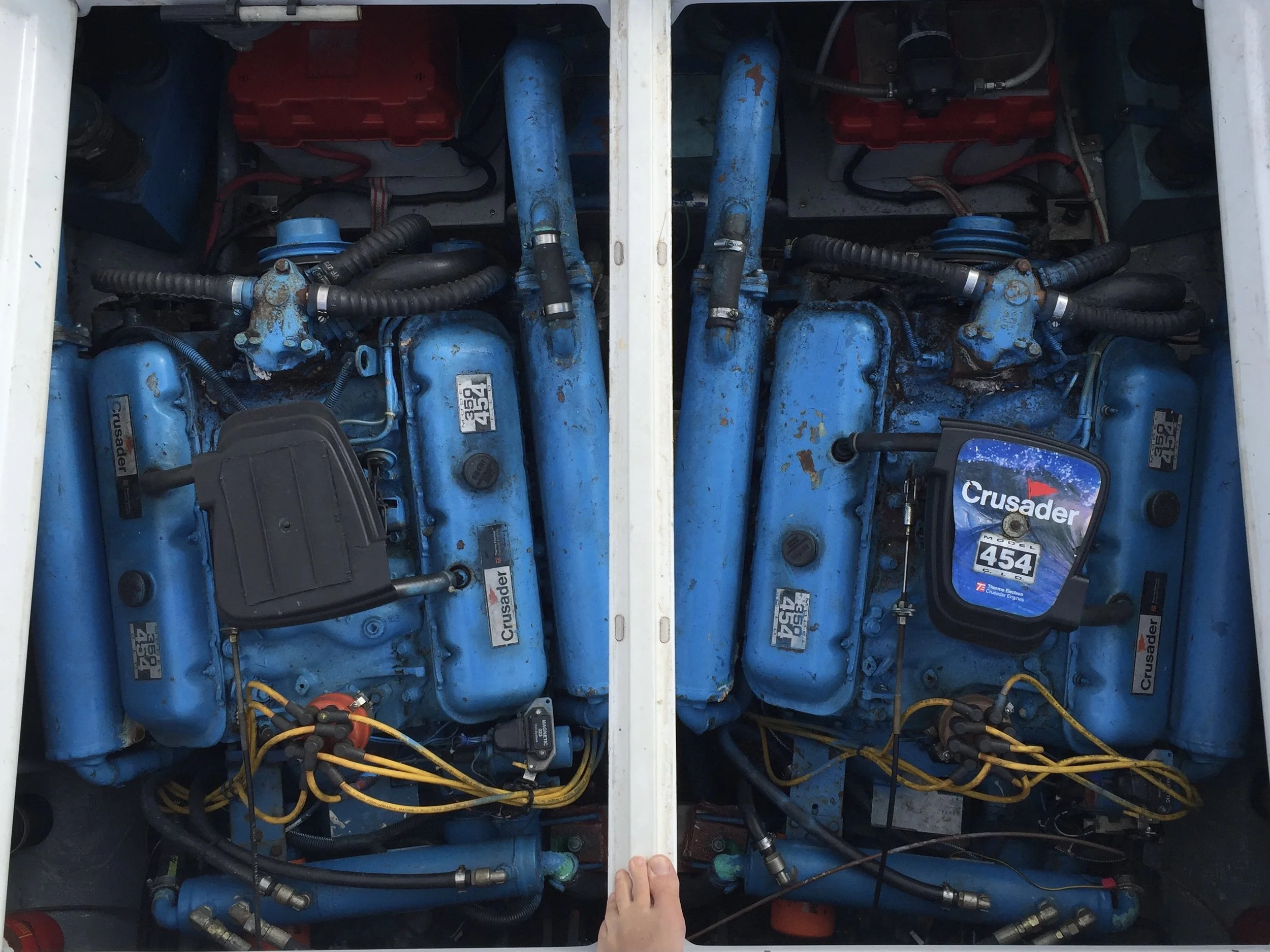 Two blue boat motors labeled 'Crusader' with yellow wiring and various hoses, positioned side by side in a boat engine compartment.