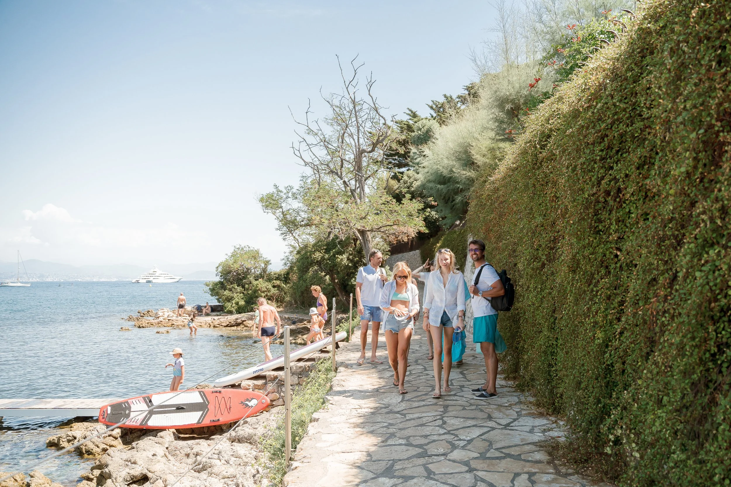 Group of people walking along a coastal path near the water, with children playing by the rocks and boats anchored in the distance on a sunny day.