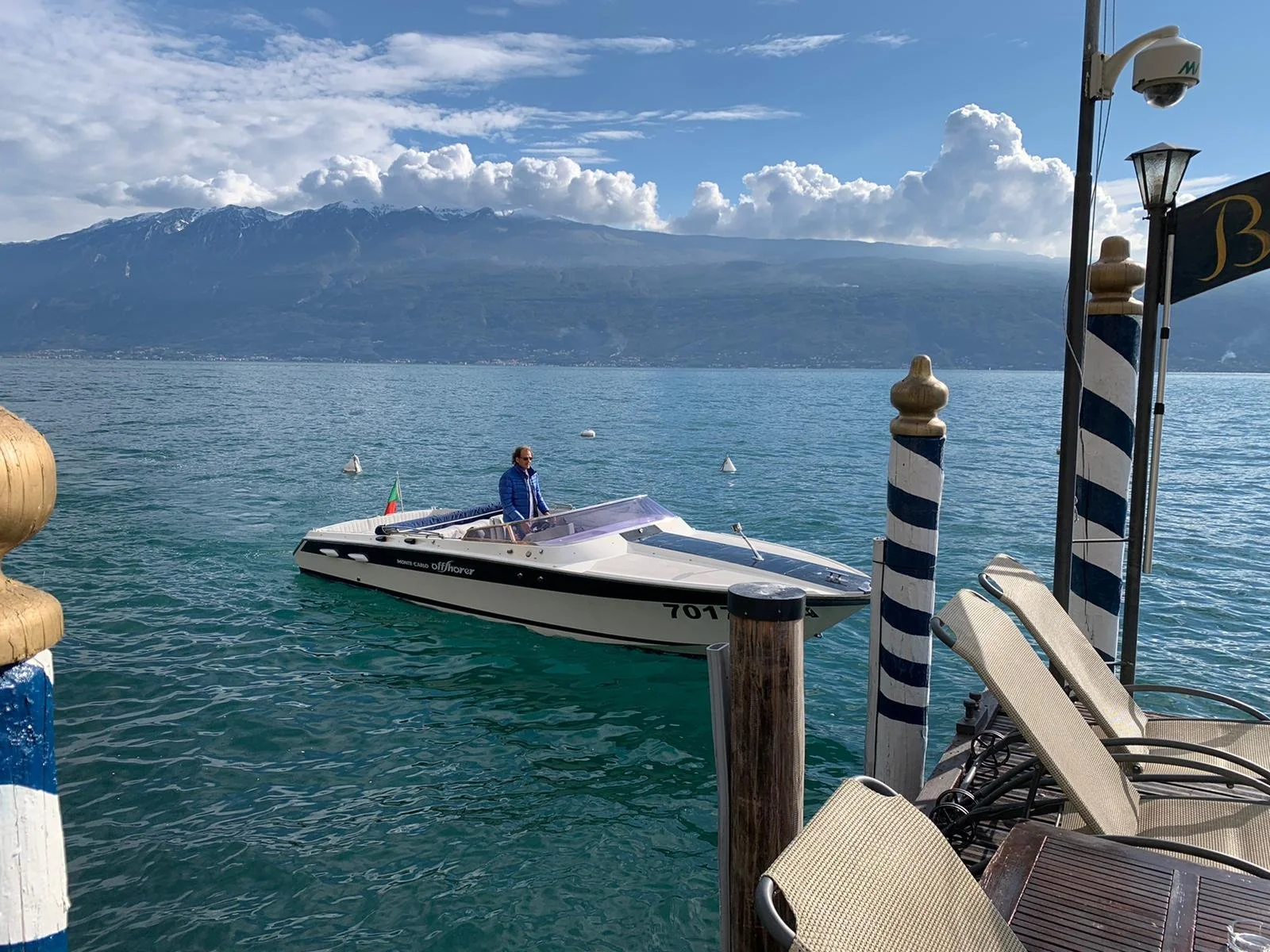 A man standing on a white, sleek motorboat in a large body of water, with mountains and partly cloudy sky in the background. The scene is viewed from a dock with striped posts, chairs, and a lamppost.