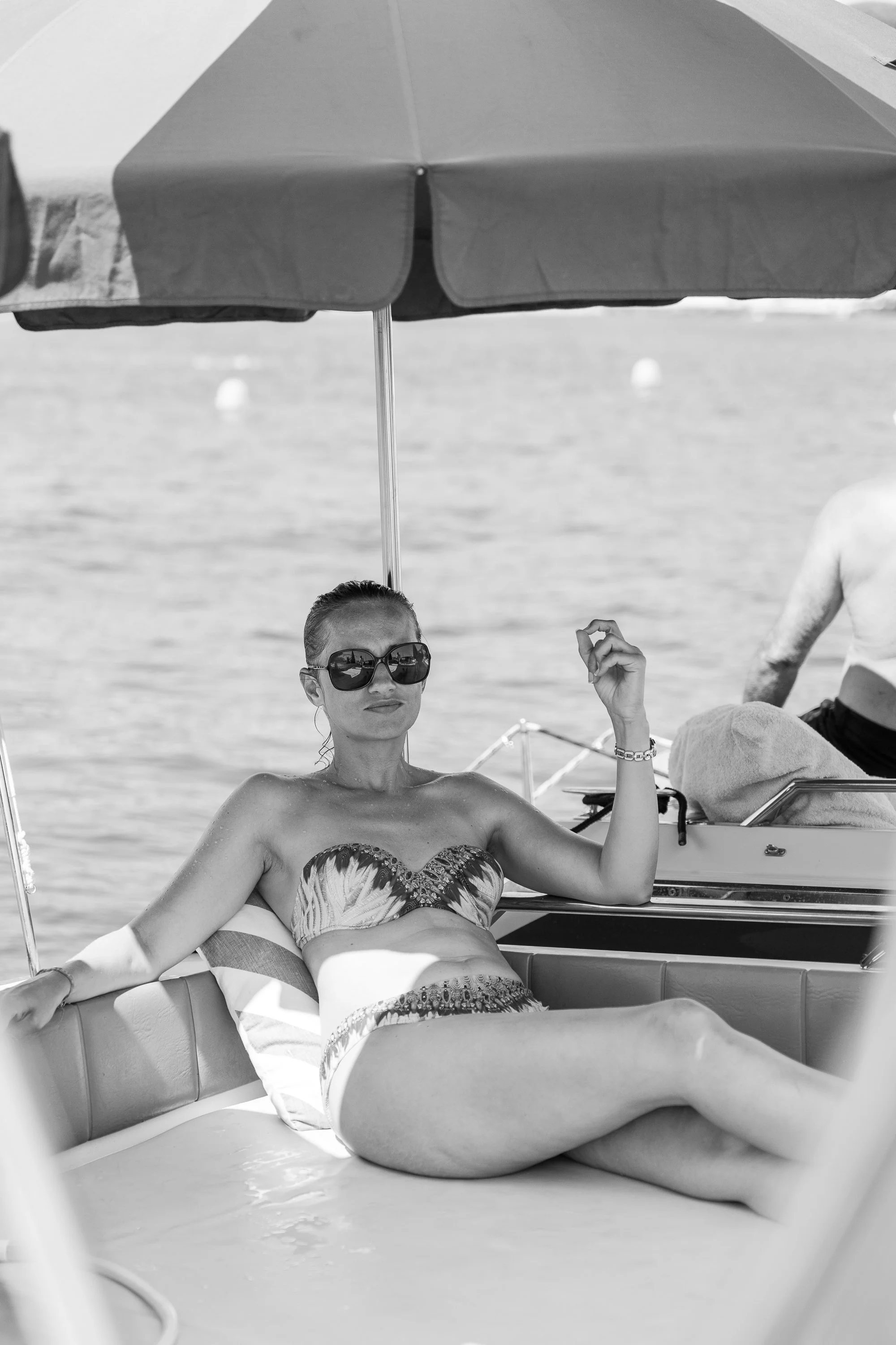 A woman relaxing on a boat wearing sunglasses and a swimsuit, sitting under a large umbrella with water in the background.
